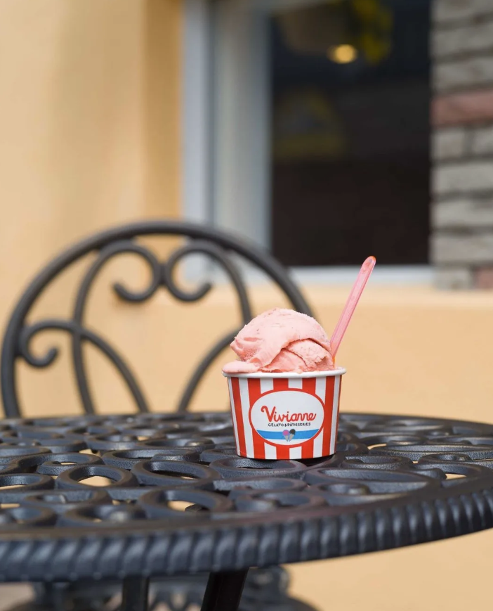 A cup of pink ice cream on a black metal outdoor table with a decorative backrest, outside a building with yellow walls and a window.