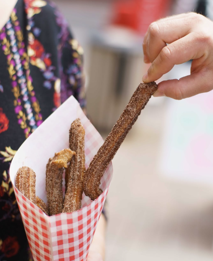 Person holding a red checkered paper tray with multiple churros coated in cinnamon sugar, one churro being pulled out.