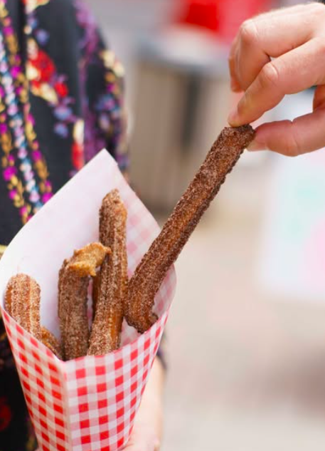 Person holding a mozzarella stick in a red and white checkered paper cone of fried churros.