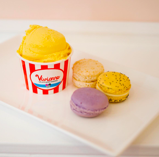 A cup of yellow lemon sorbet alongside three macarons in light purple, yellow with black specks, and beige colors on a white plate.