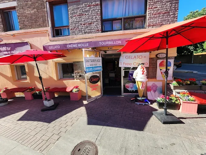 Ice cream shop with red umbrellas, pink flower pots, and a sign advertising gelato and frozen treats outside the entrance.