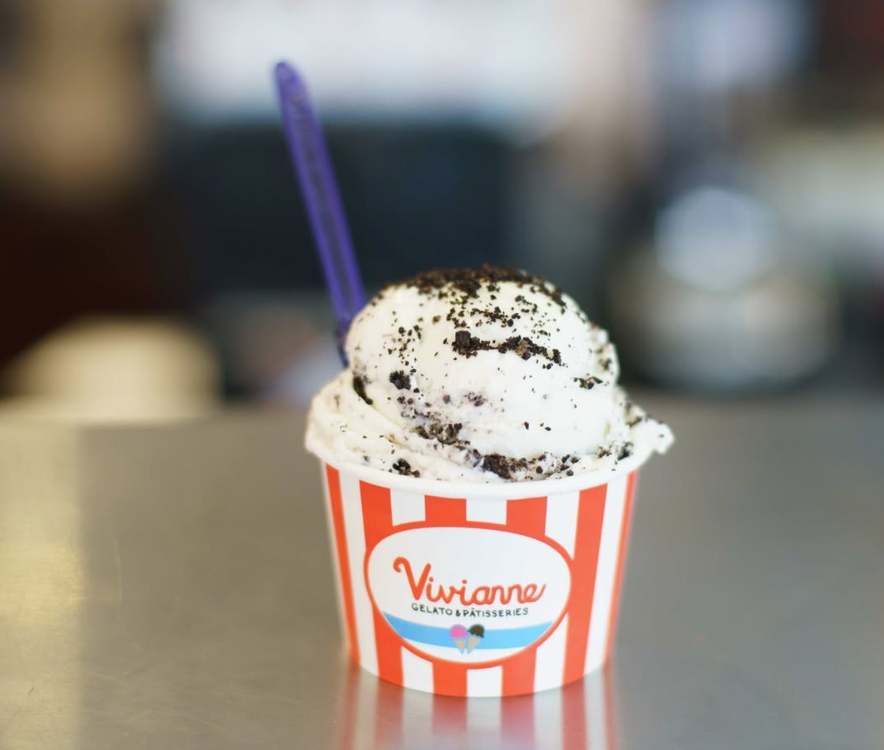 A cup of cookies and cream ice cream with crushed cookie pieces in a red and white striped paper cup. The cup has a Vivianne gelato & patisserie logo. A purple spoon is sticking out of the ice cream.