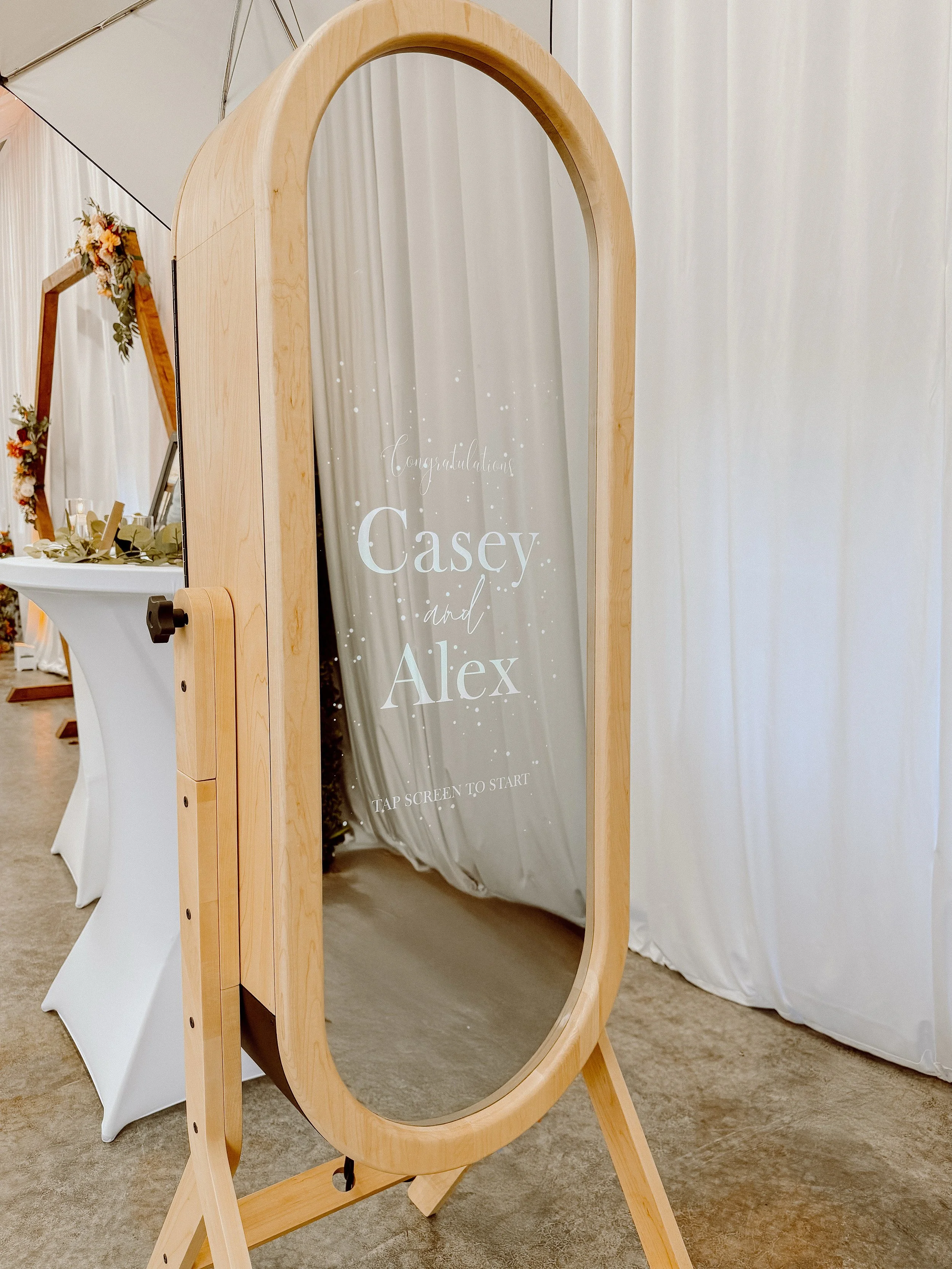 Full-length wooden-framed mirror with a digital message displaying congratulations to Casey and Alex, inviting guests to tap the screen to start, set against white curtains and a decorated table.