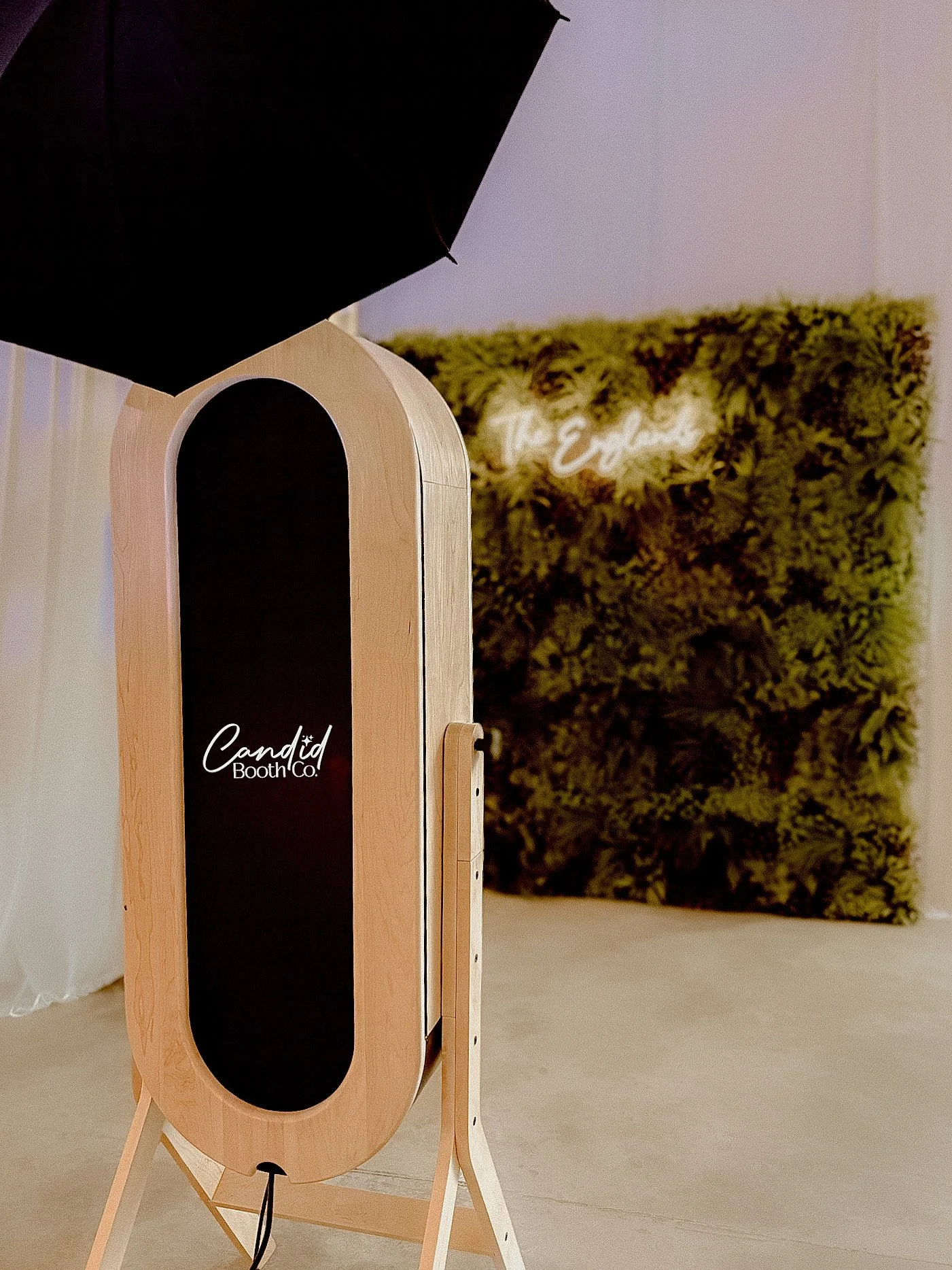 A photo booth with a black chalkboard front and wooden frame, positioned indoors, with a backdrop of green moss wall and illuminated sign.
