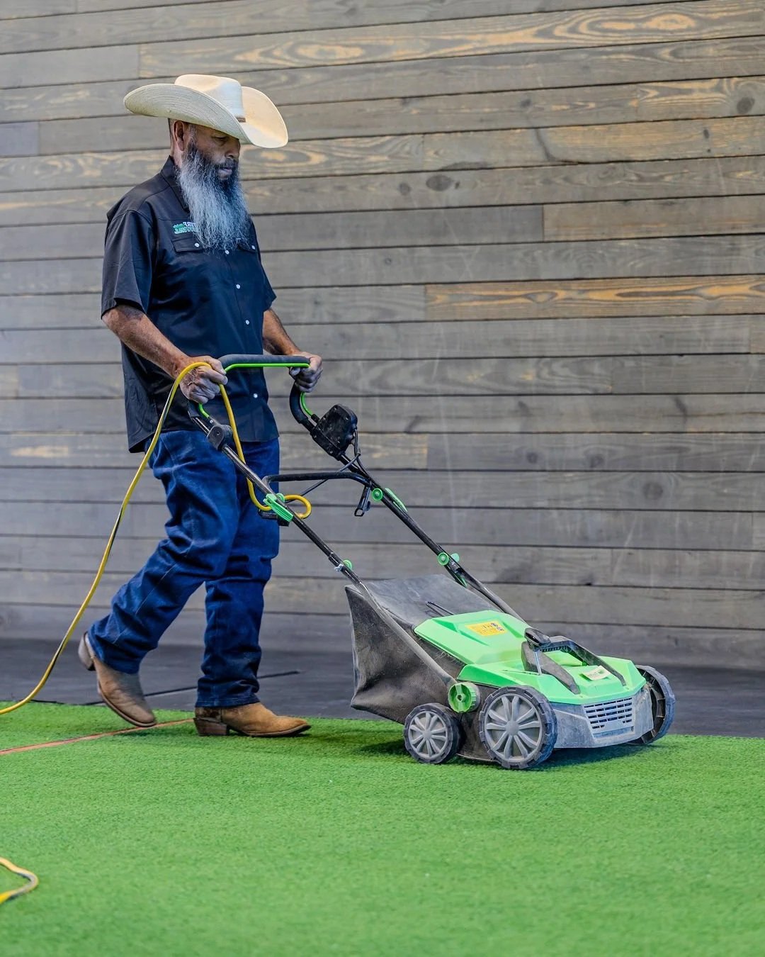 A man with a long gray beard, wearing a cowboy hat, black shirt, blue jeans, and brown boots, is using a green and black walk-behind lawn mower on a small patch of artificial grass with a wooden wall in the background.