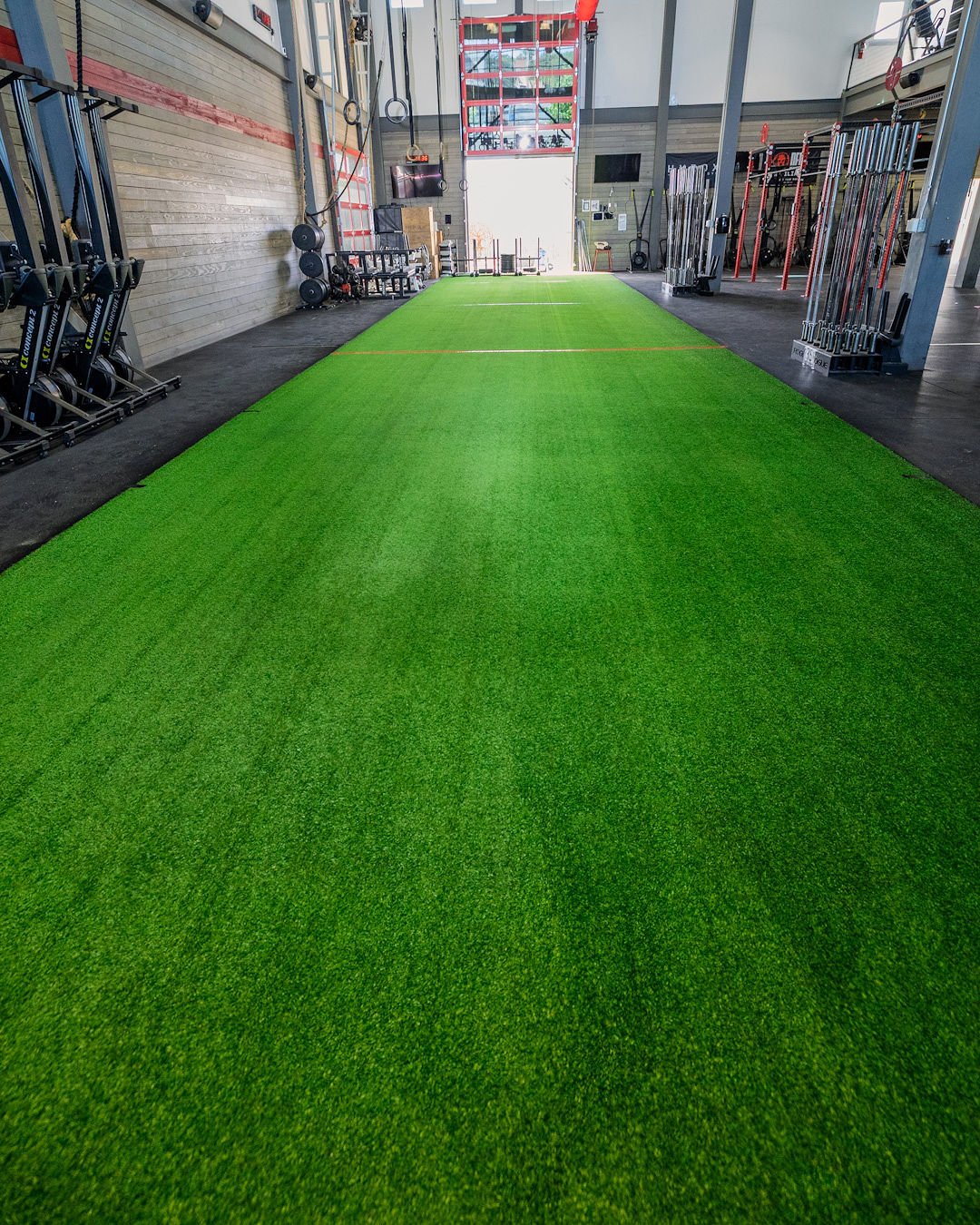 Indoor gym with a green turf workout area, gym equipment on both sides, and large windows at the back letting in natural light.