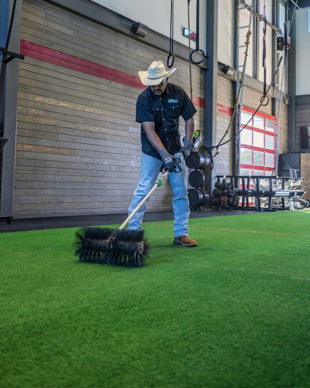 A man wearing a cowboy hat, sunglasses, a black shirt, and light blue jeans is using a floor sweeper on a green indoor sports surface. The background features gym equipment like weights and gymnastic rings.
