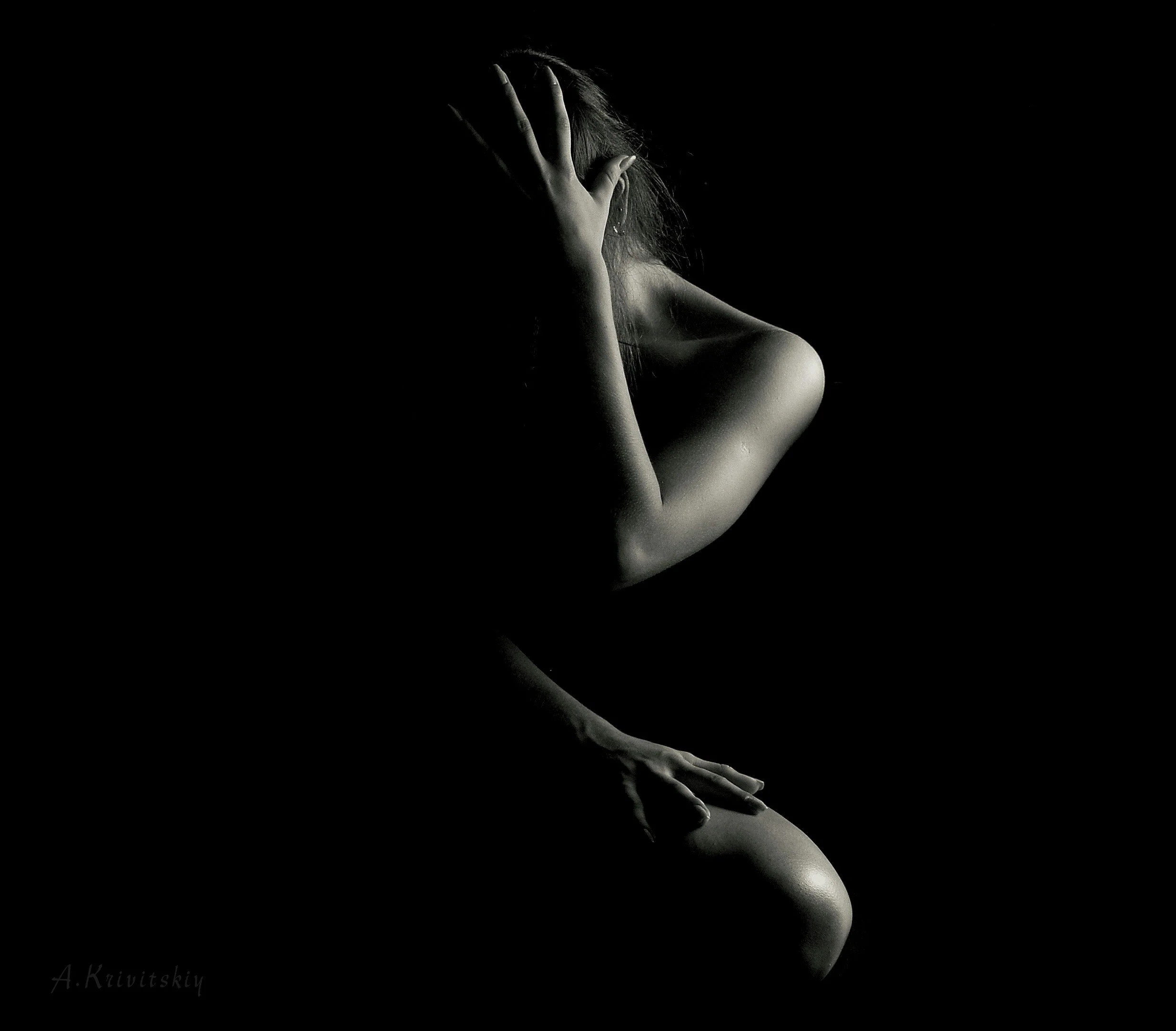 A black and white photograph of a nude woman in a contemplative pose, with her head resting on her hand, highlighting her shoulder and arm against a dark background.