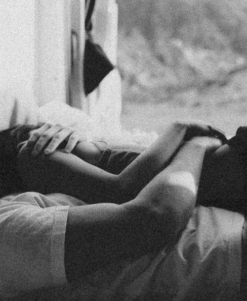 A person lying on a bed holding hands with another person, with a window and outdoors visible in the background.