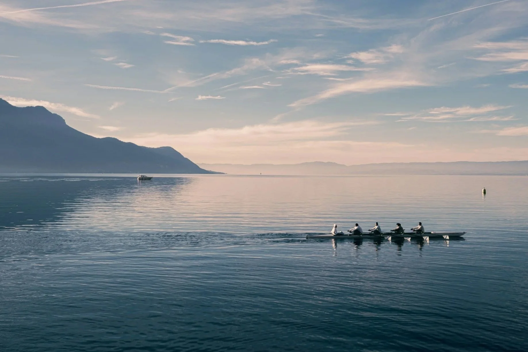 A team of five rowers in a racing shell on a calm body of water during the daytime, with mountains and scattered clouds in the background.