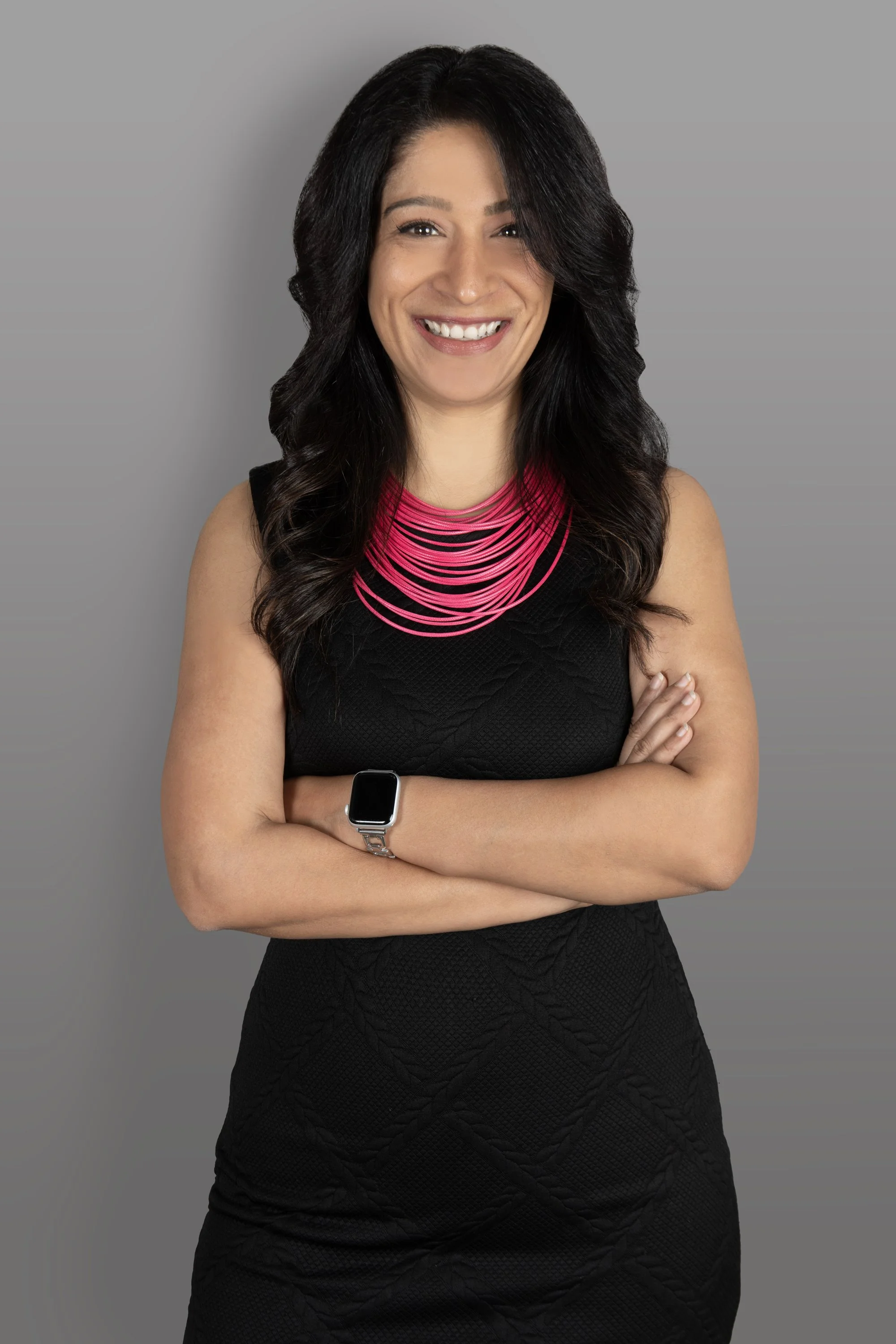 A woman with long dark wavy hair smiling, wearing a black sleeveless dress, pink layered necklace, and a smartwatch with crossed arms against a gray background.