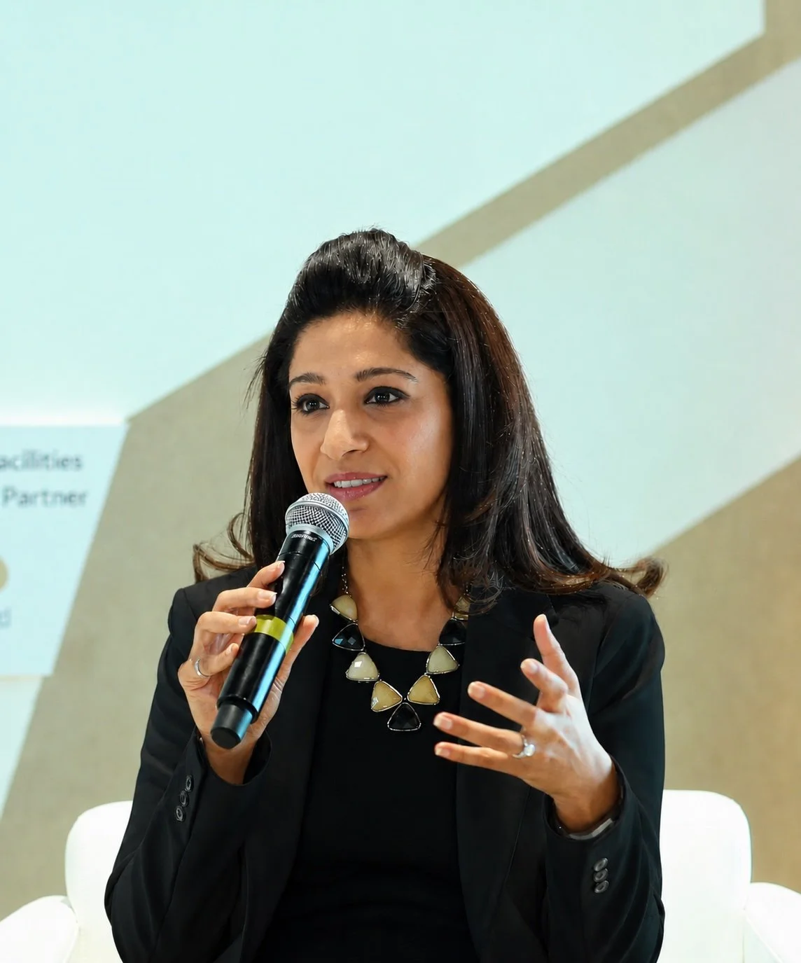 A woman with dark hair, wearing a black blazer and necklace, speaking into a microphone at an event or conference.