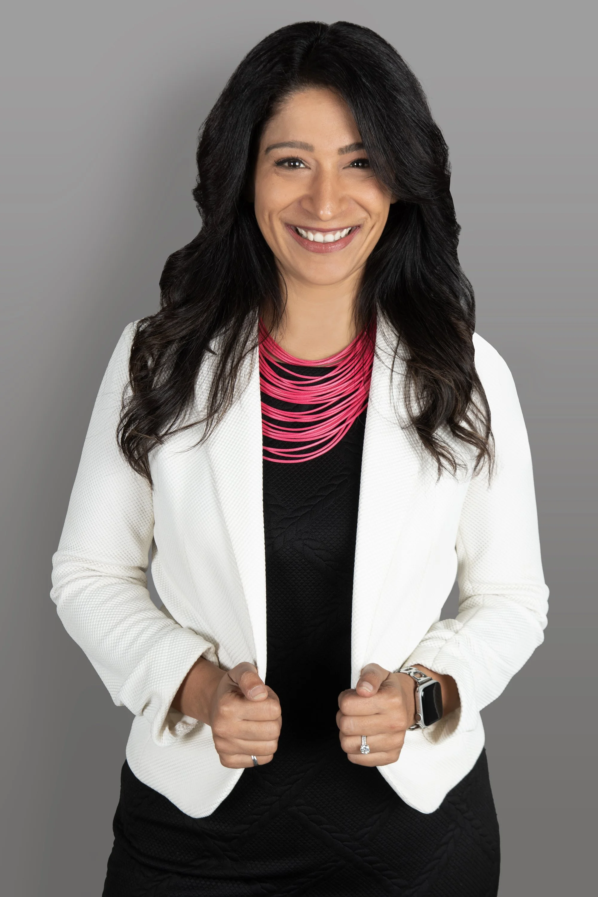 A woman with long black hair, smiling, wearing a white blazer, black top, a pink necklace, a smartwatch, and a ring, against a gray background.
