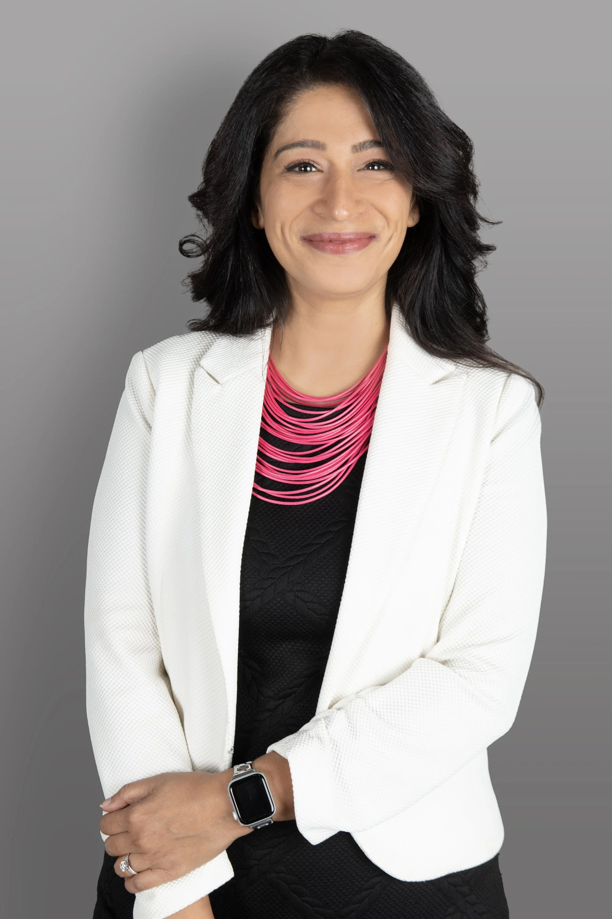 A woman with shoulder-length black hair, smiling, wearing a white blazer, and a pink and black striped top, standing with arms crossed and wearing a smartwatch on her left wrist against a gray background.