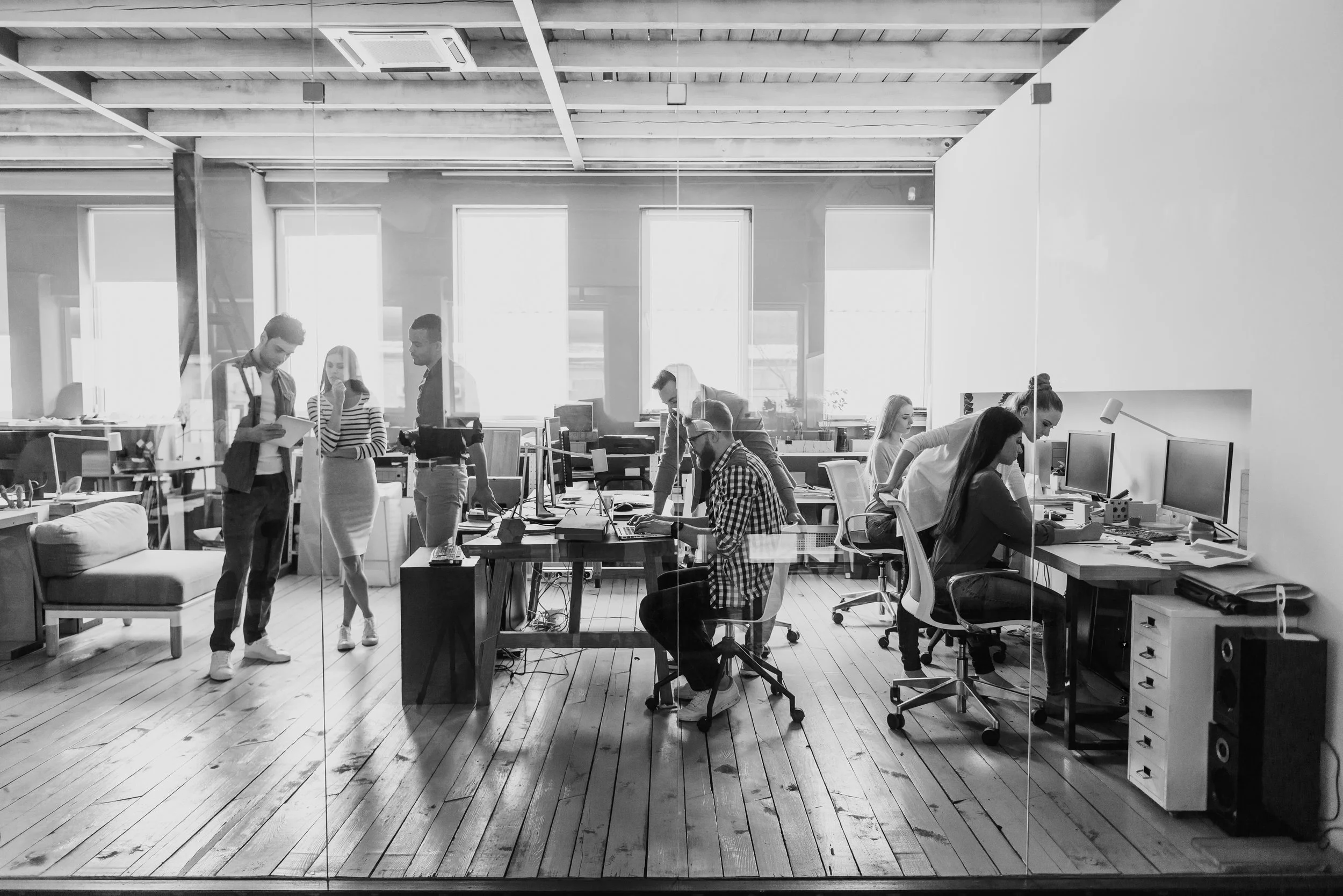 Vista de una oficina moderna con varias personas trabajando y colaborando, algunos sentados frente a computadoras y otros de pie, en un ambiente luminoso y organizado.