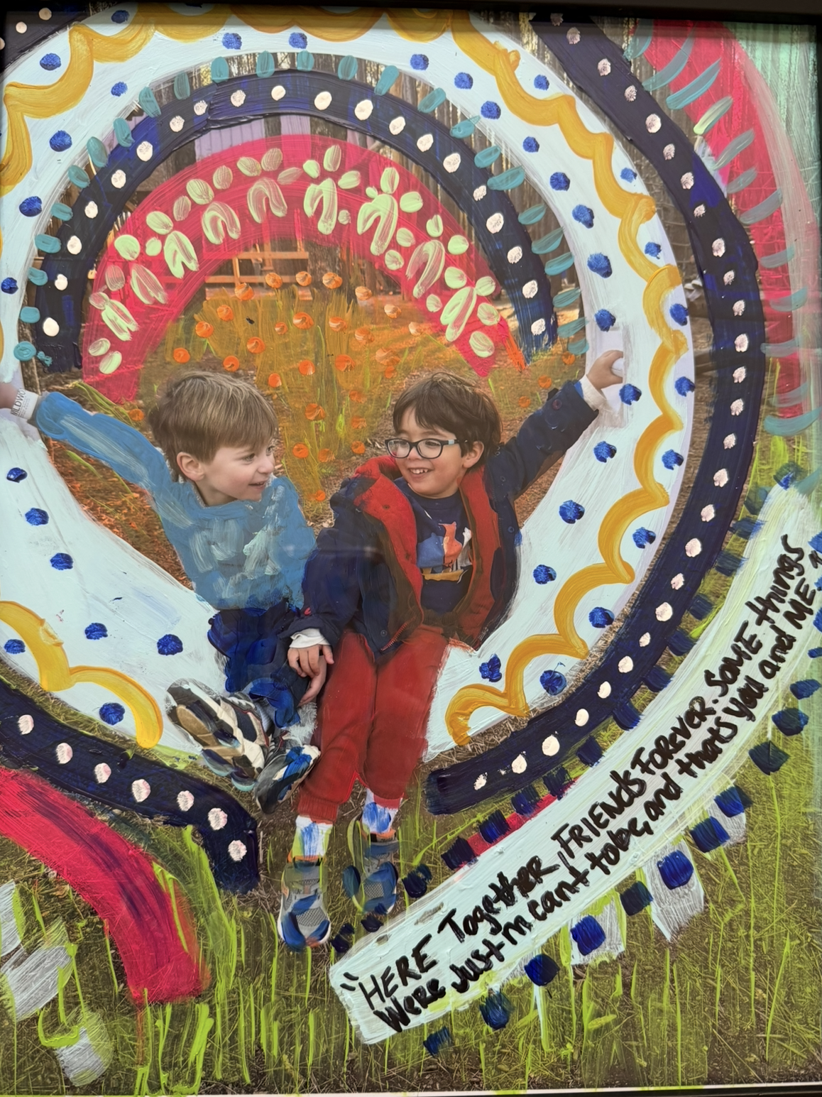 Photo of two young boys playing outdoors, surrounded by colorful, painted designs including hearts and swirls. The boy on the left is smiling and wearing a blue jacket, while the boy on the right is smiling broadly, wearing glasses, a dark blue jacket, and red pants, with his arm raised. The background features grassy ground and the scene is decorated with vibrant, painted patterns and a quote that reads, "Here together, forever soul hung, we're just innocent and I want you are me."