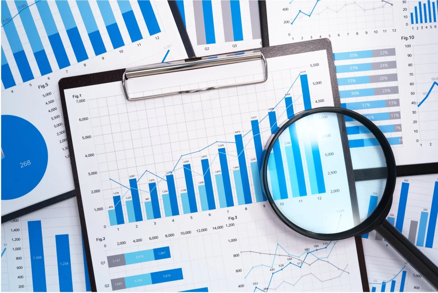 Multiple business charts and graphs with blue and gray color schemes on papers and screens, a magnifying glass highlighting one chart, and a clipboard holding a sheet with a line graph.