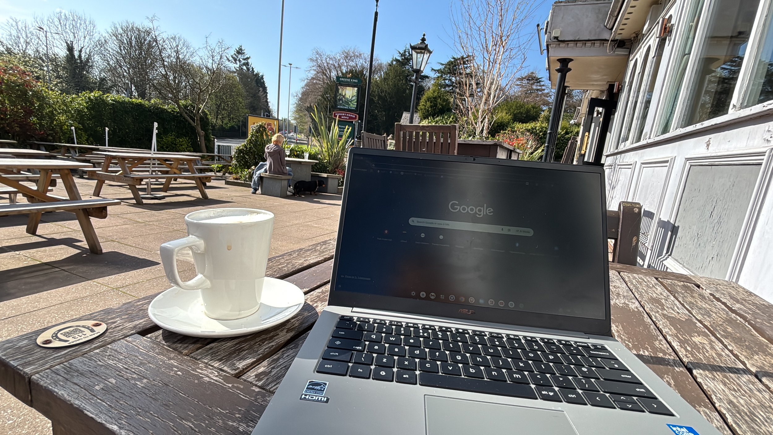 A table with a laptop and a white coffee mug outside on a wooden picnic table in a sunny outdoor setting. In the background, a person sitting on a bench next to a small dog, trees, and a street with signs and street lamps.