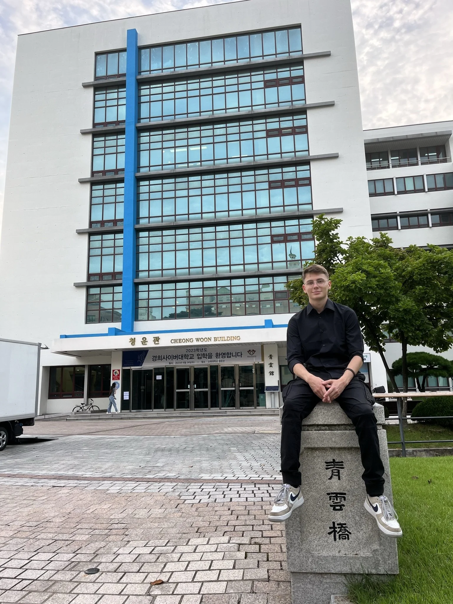 A young man in black clothing sits on a stone sculpture with Asian characters carved into it, in front of a modern multi-story building with large glass windows and a white facade. There are trees and a cloudy sky in the background.