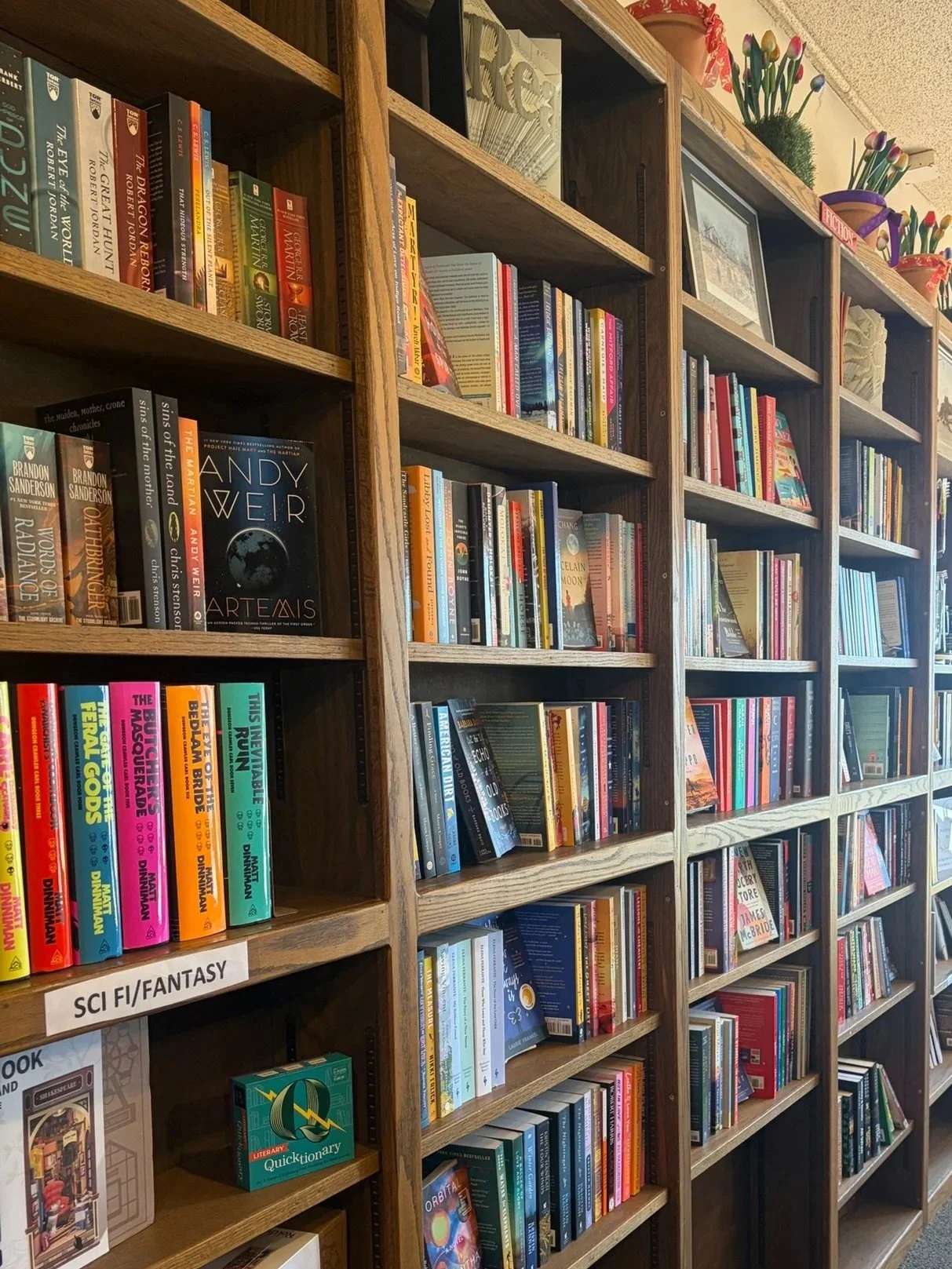 Wooden bookshelves filled with various books, including fiction, science fiction and fantasy titles, in a bookstore. Potted plants with colorful flowers are placed on top.