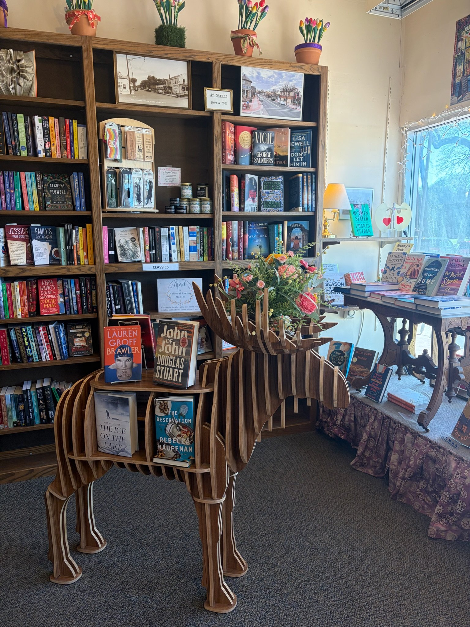 Interior of a bookstore with a wooden horse-shaped bookshelf filled with books, a table with more books and a flower arrangement, and a bookshelf with framed photos and potted plants on top. There is natural light coming through a window.