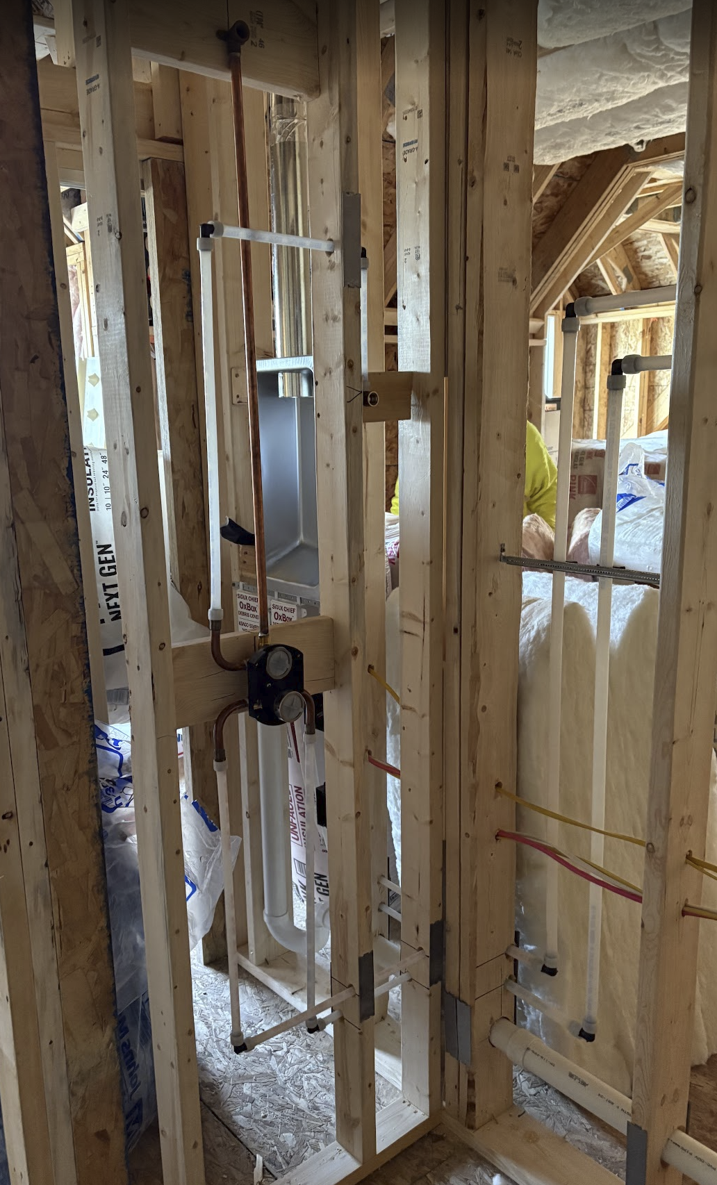 Interior framing of a building under construction with plumbing pipes, electrical wiring, and insulation visible.