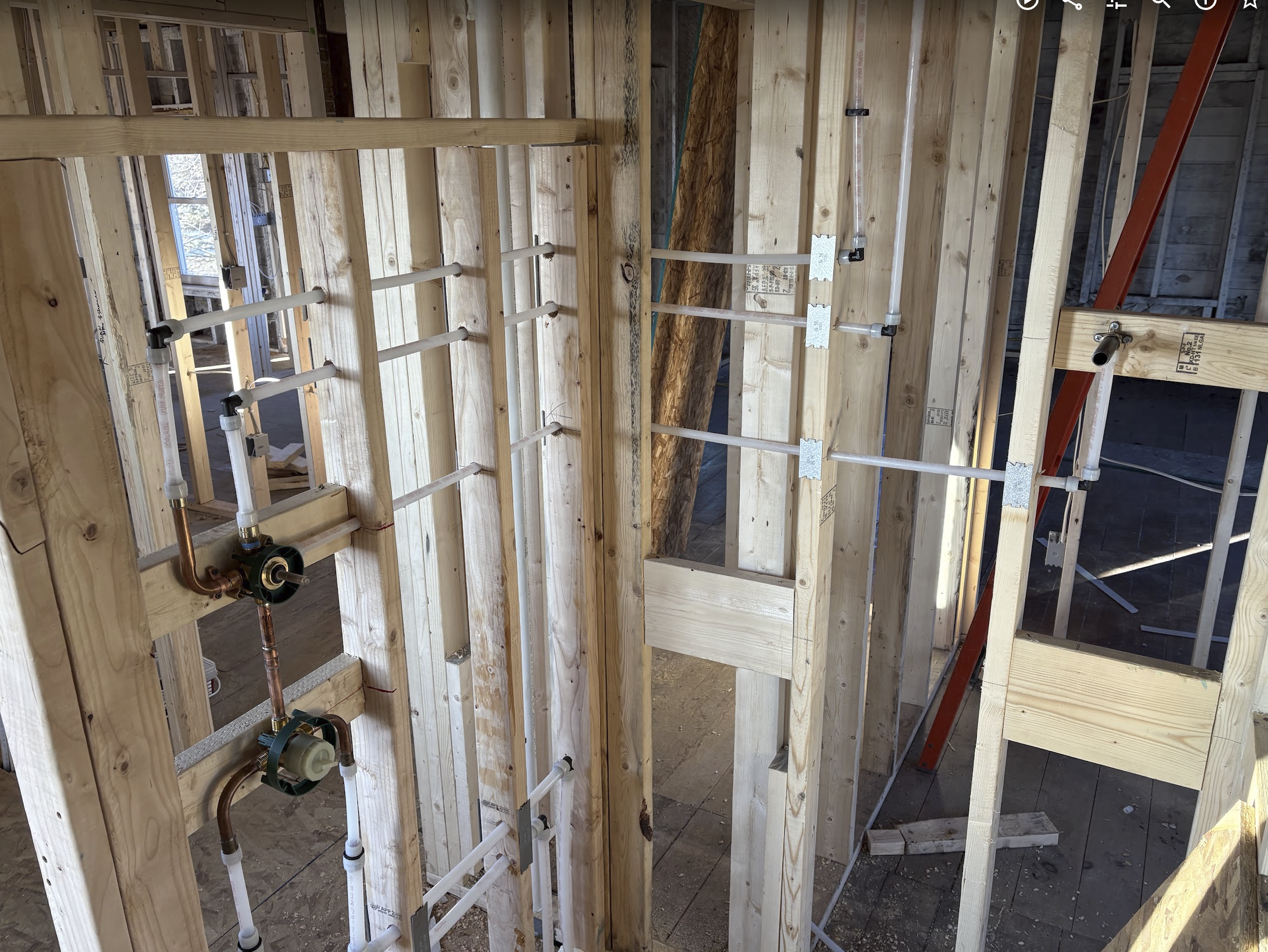 Interior view of a residential construction site with exposed wooden framing, plumbing pipes, and electrical wiring in the early stages of building the walls.