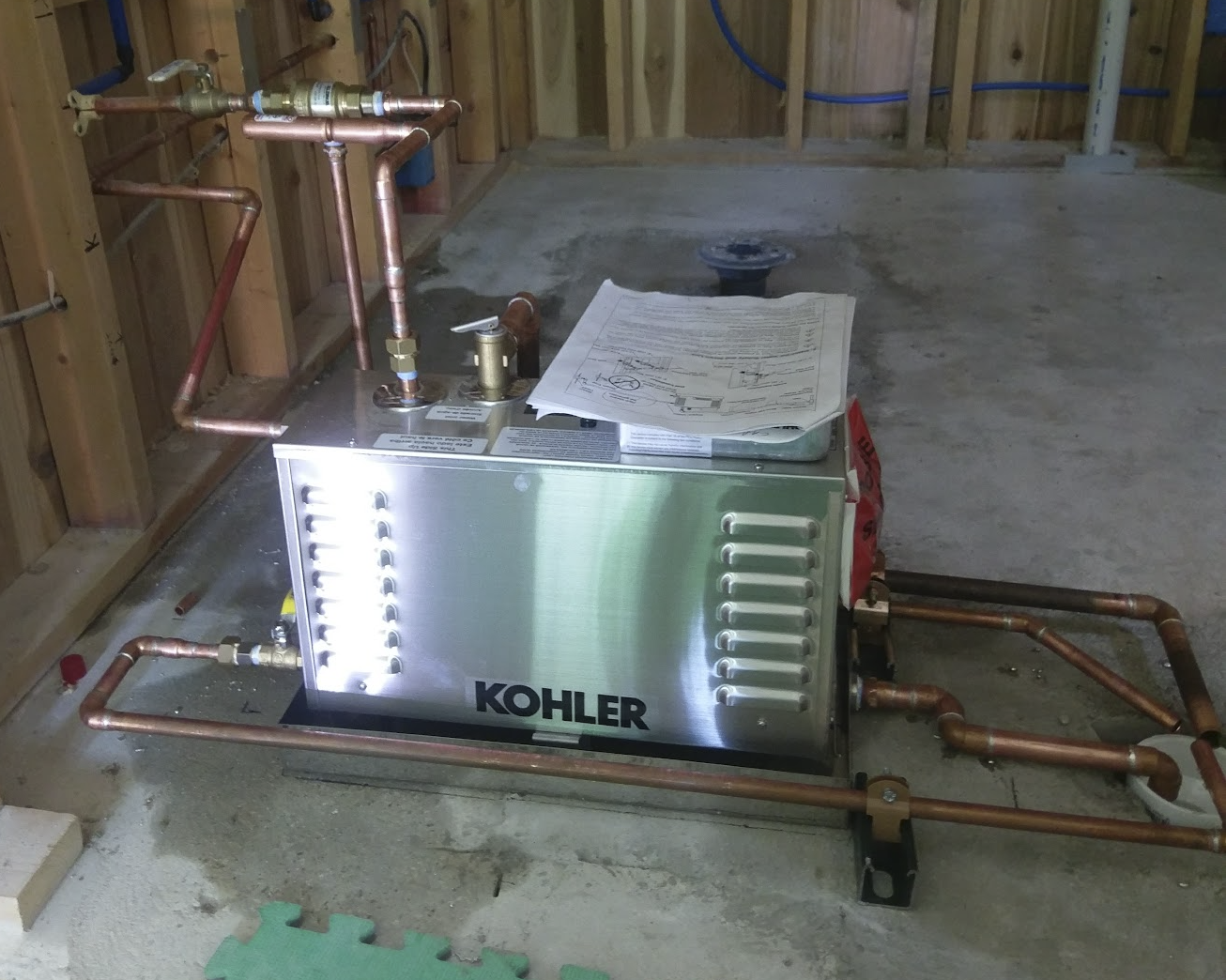 A Kohler HVAC water heater installation in a room under construction with exposed wooden studs, plumbing pipes, and a concrete floor.