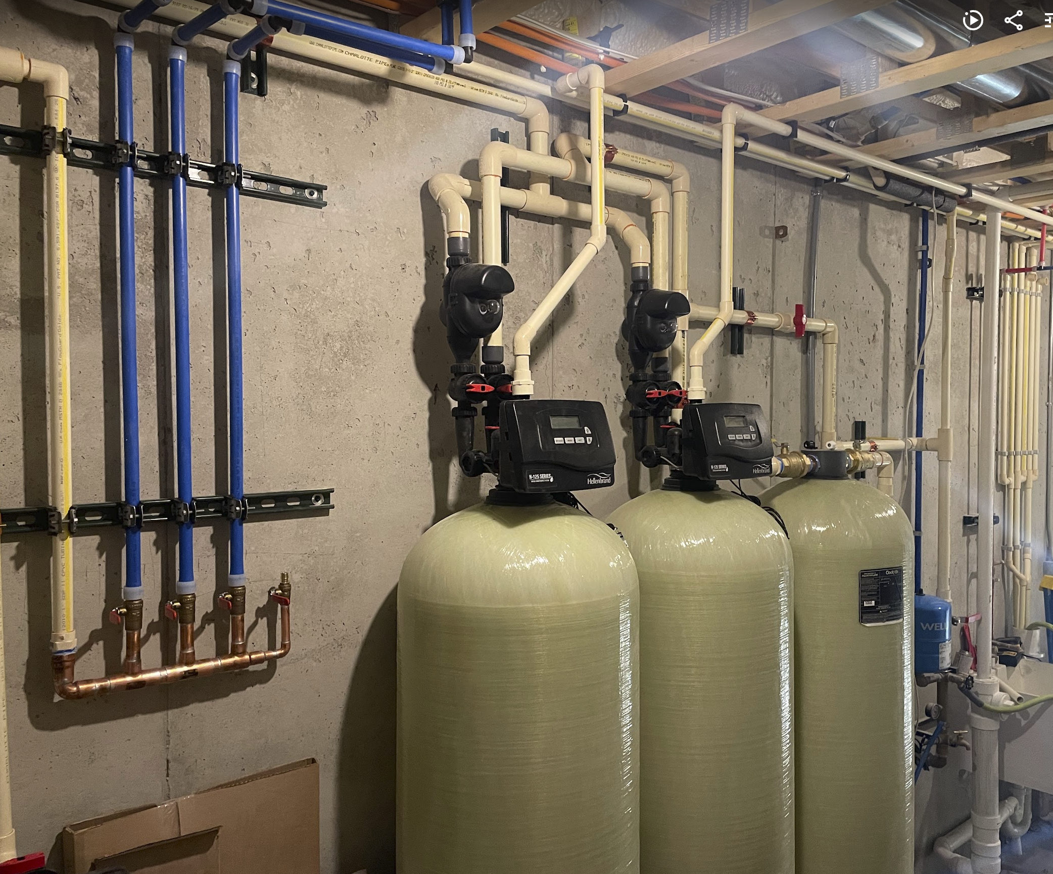 A basement utility room with water filtration or softening systems and plumbing pipes mounted on the concrete wall.