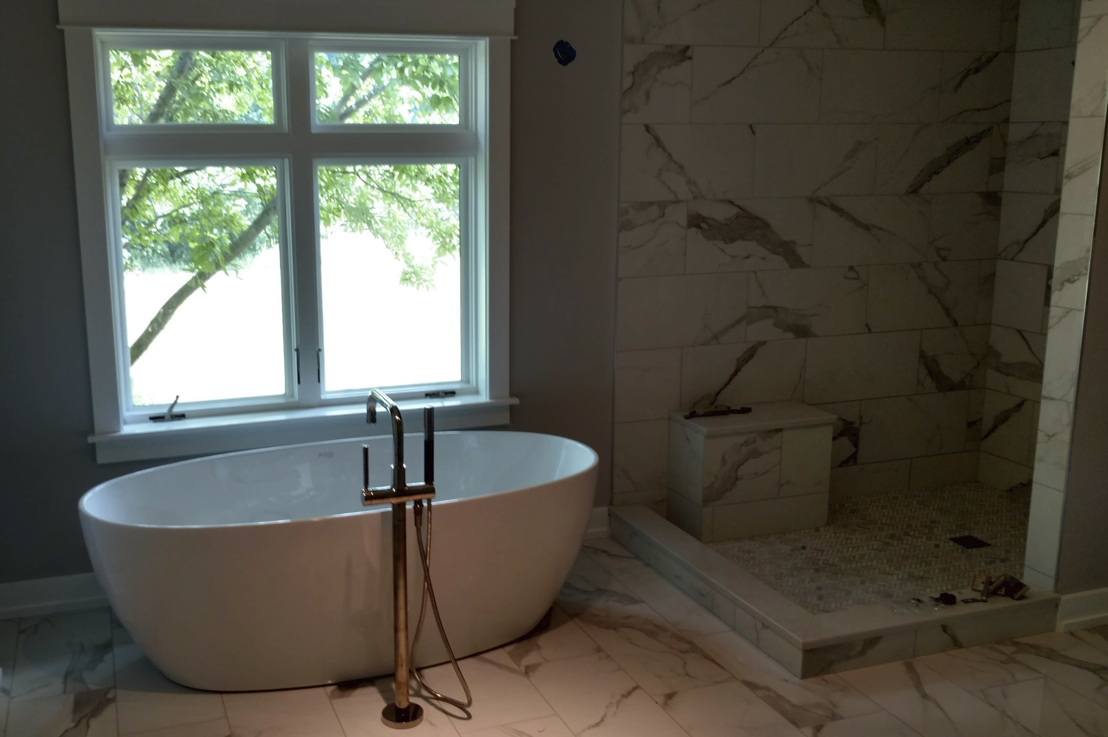 A modern bathroom with a freestanding white bathtub under a large window with green trees outside, and a marble-tiled shower area to the right.