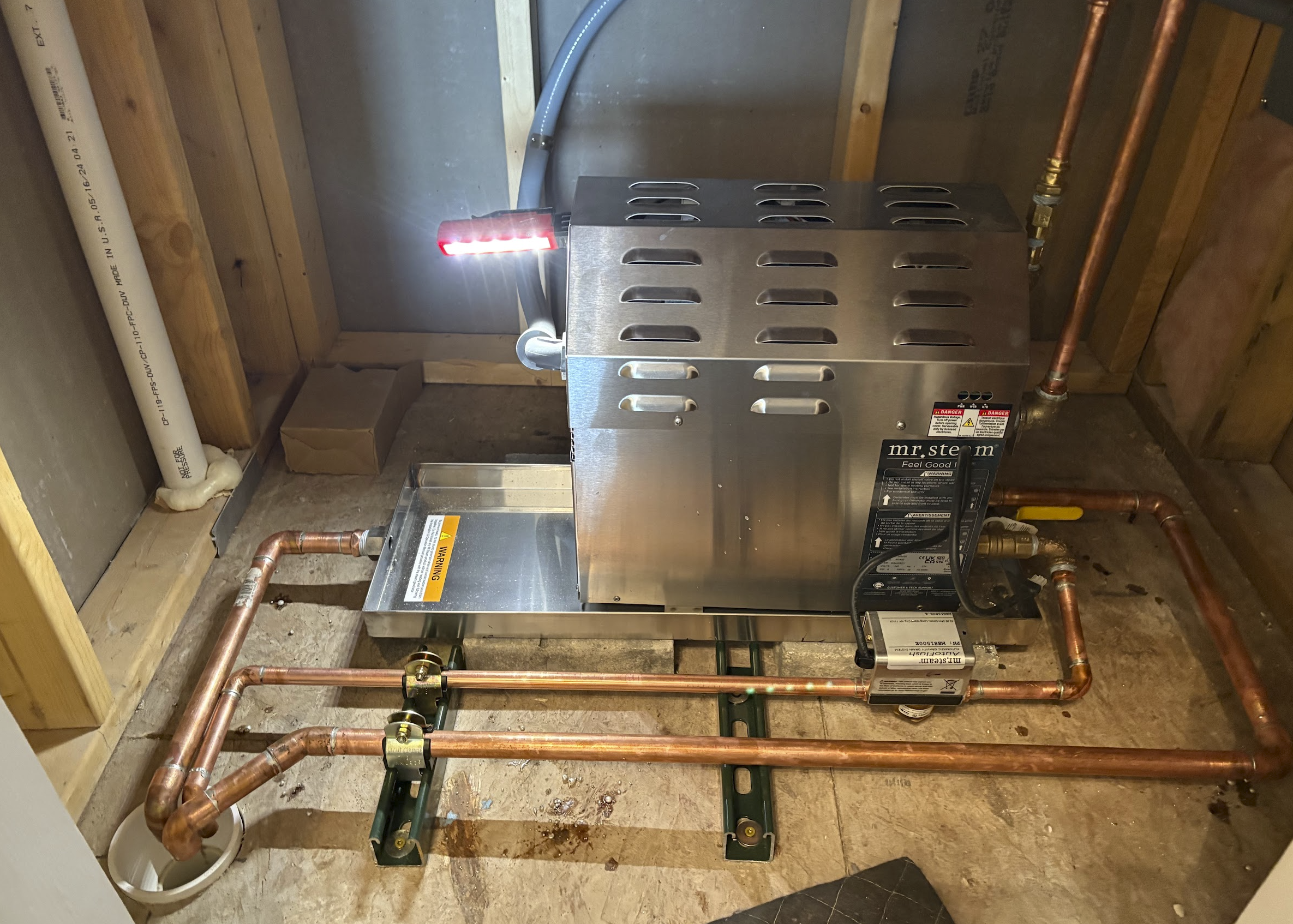 A water or heating system with copper pipes, a metallic Mr. Steem unit, and a red LED indicator, installed in a wooden framed basement or utility room.