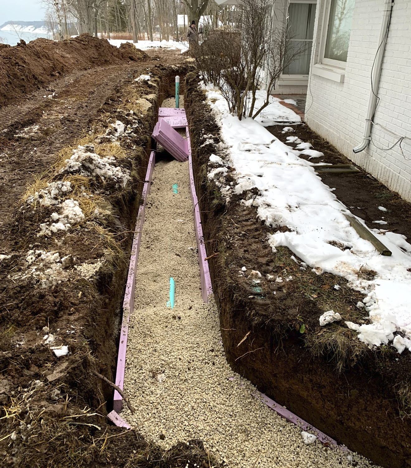 Construction site with a narrow trench filled with gravel, pink framing, and partially buried purple insulation, next to a white house with snow on the ground and a partially visible person in the background.