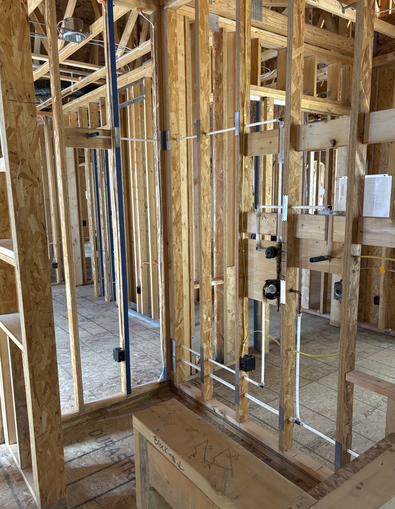 Interior view of a building under construction showing exposed wooden framing, pipes, electrical wiring, and unfinished walls.