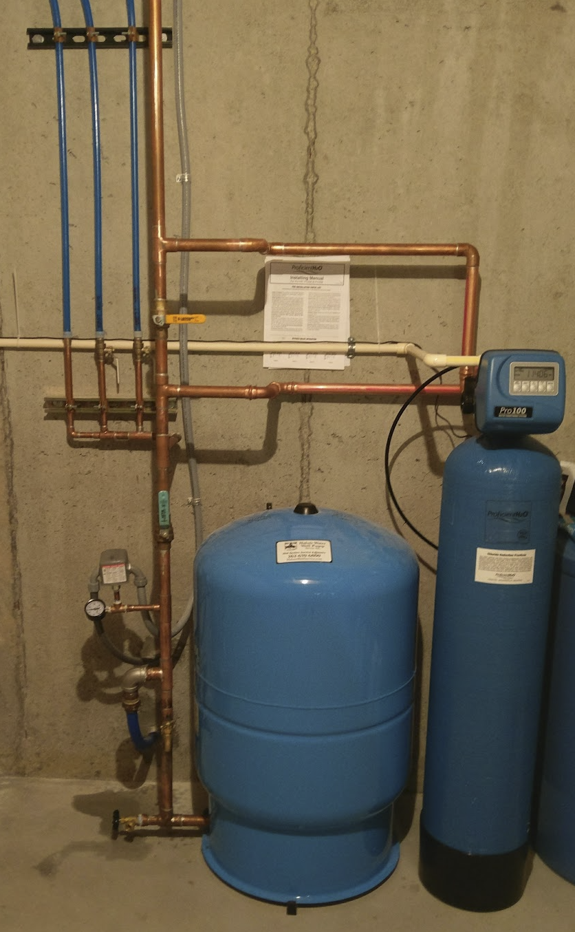 A water filtration and softening system with copper pipes, a large blue tank, a control valve, and a filtration unit against a concrete wall.