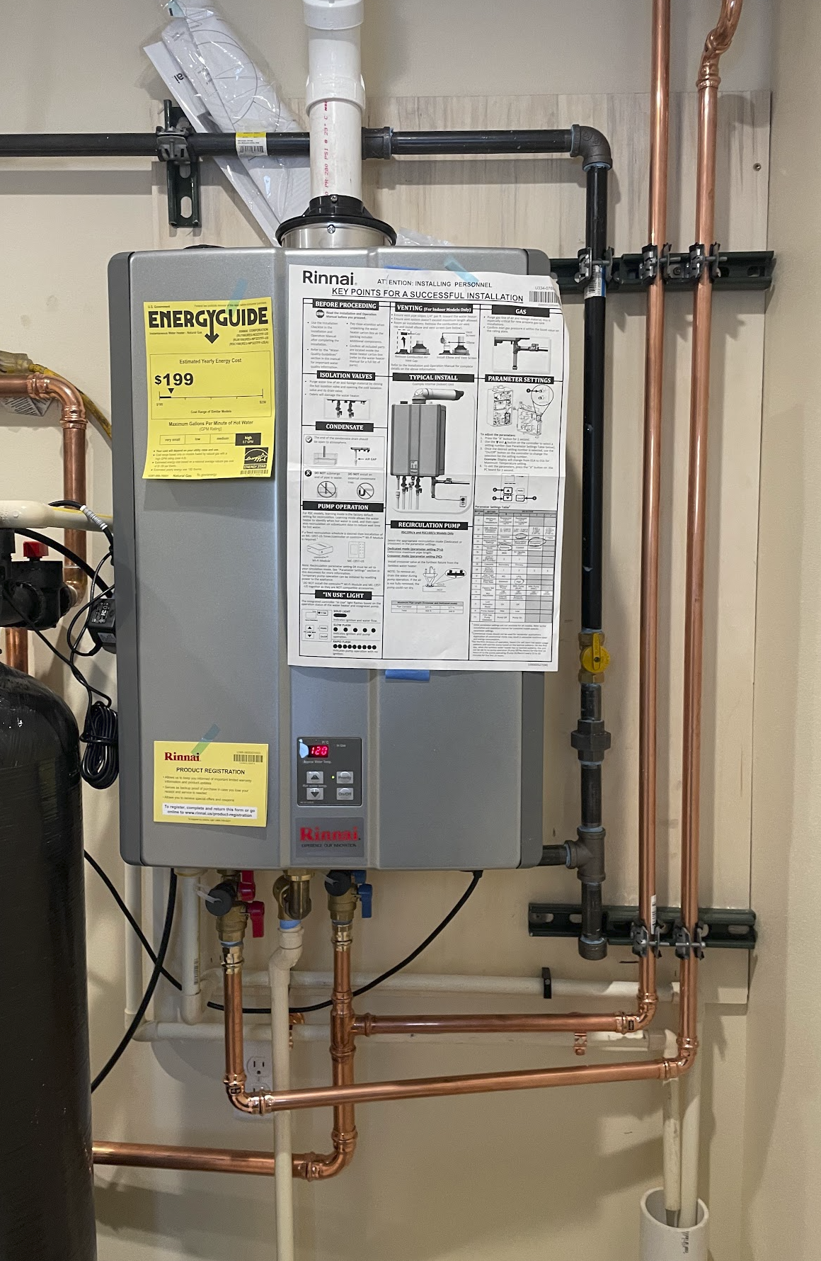 A water heater unit mounted on a wall with various pipes connected to it, including copper and black pipes, and instruction labels attached.
