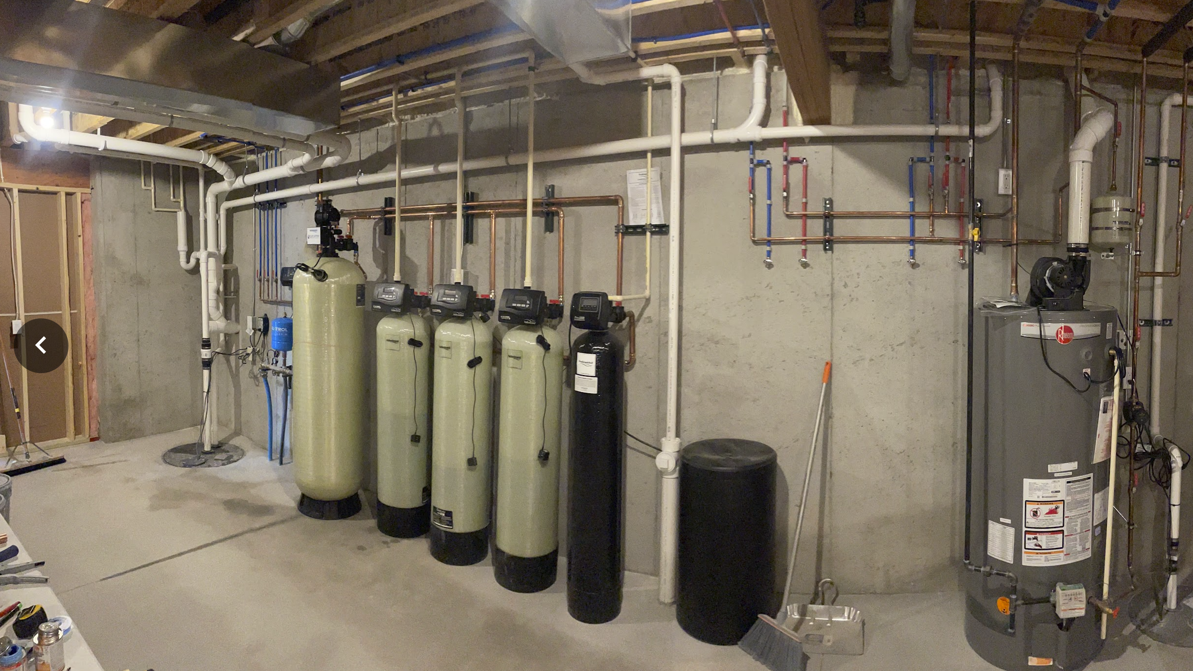 A basement water softening and filtration system with multiple tanks, pipes, and control units installed on a concrete wall.