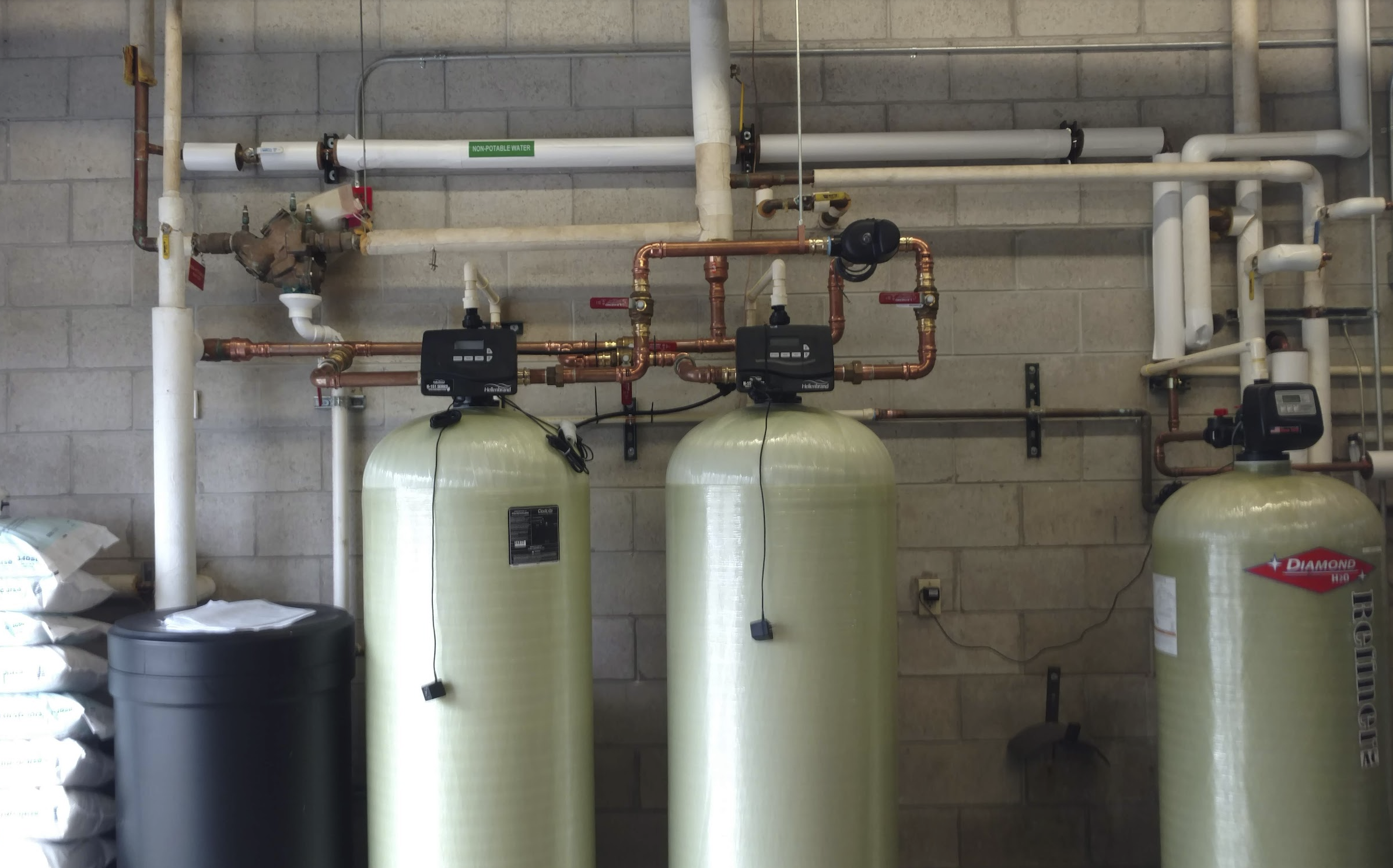 Industrial water filtration system with three large green tanks, copper pipes, and control units against a concrete block wall.
