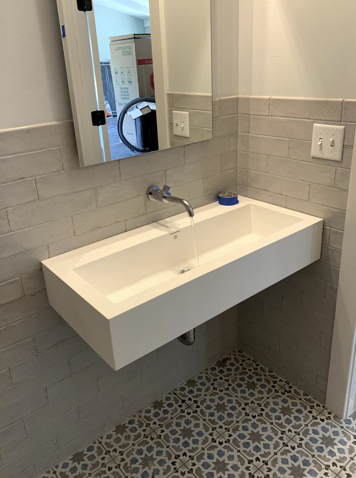 A bathroom sink with a mirror above it, a roll of blue painter's tape on the sink, and a gray tiled wall with two electrical outlets. The sink has water running from the faucet.