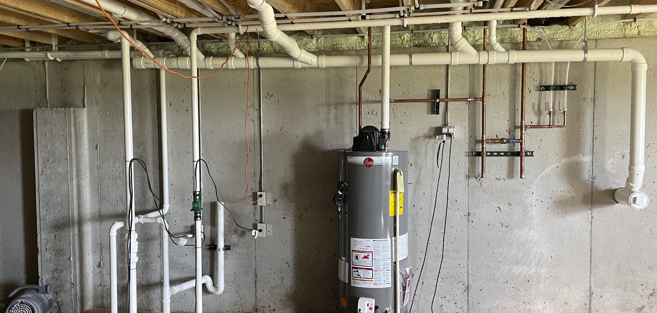 An unfinished basement wall with plumbing pipes, a water heater, and electrical wiring.