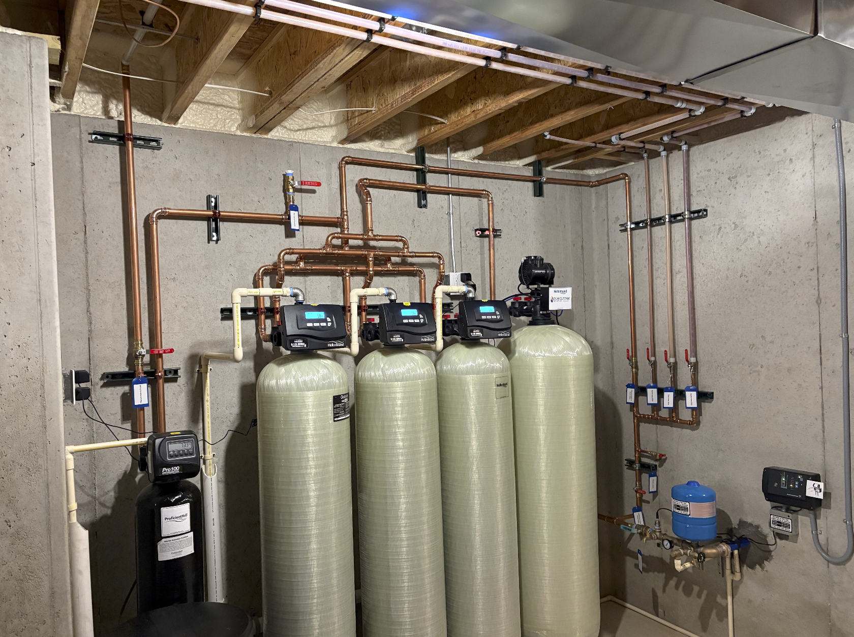 A water filtration system with multiple large green tanks, copper pipes, and control units installed against a concrete wall in a basement or utility room.