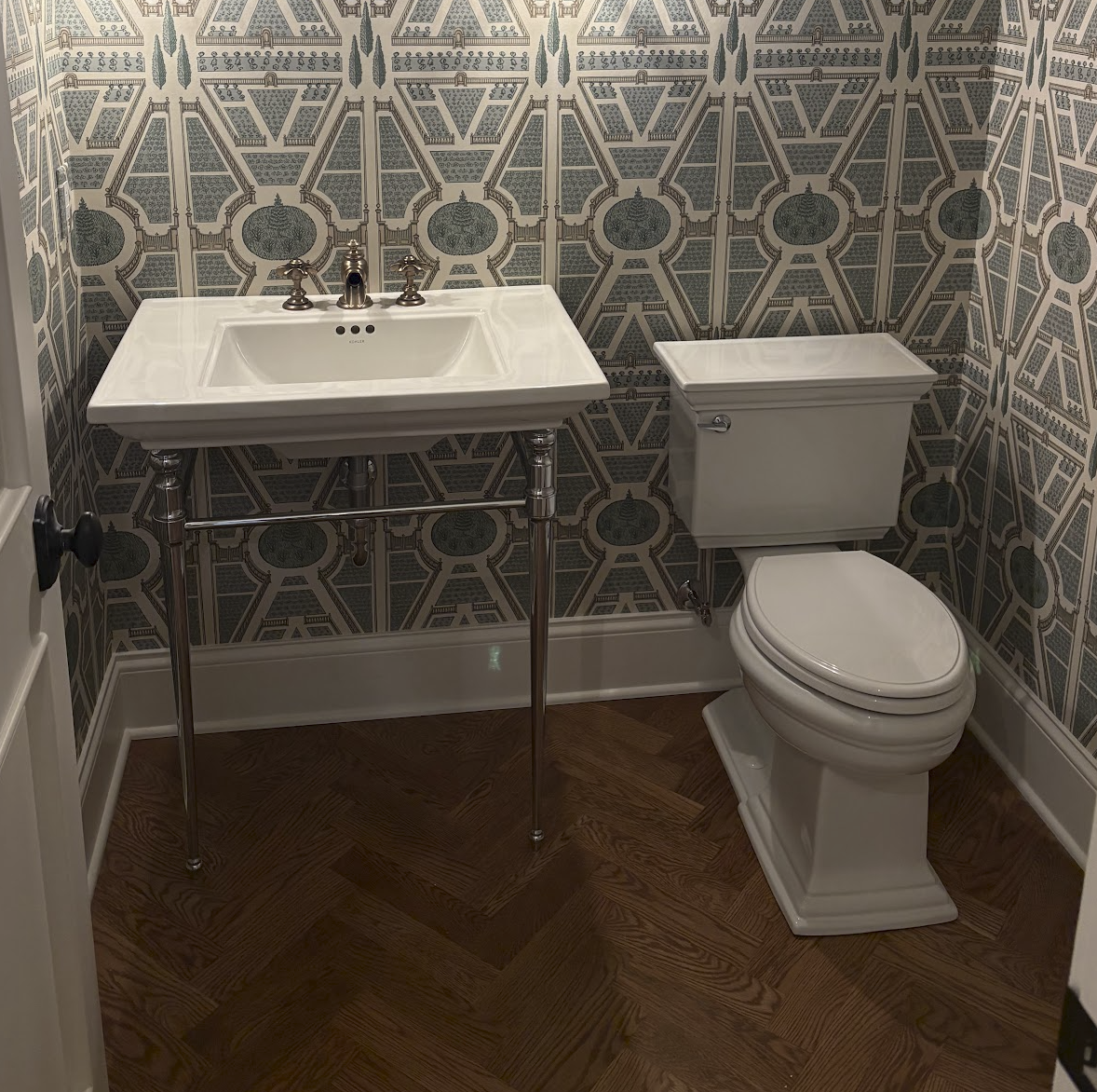 A small bathroom with vintage-style wallpaper, a white pedestal sink with chrome legs, and a toilet with a rectangular tank. The room has dark wood flooring and a white baseboard.