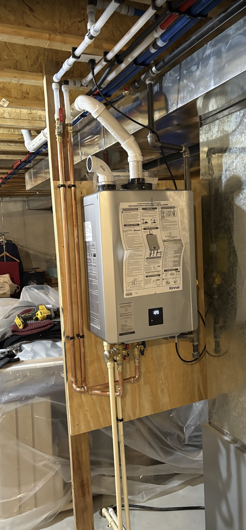 A water heater with plumbing pipes and vents in a basement or utility room, with tools and construction materials nearby.