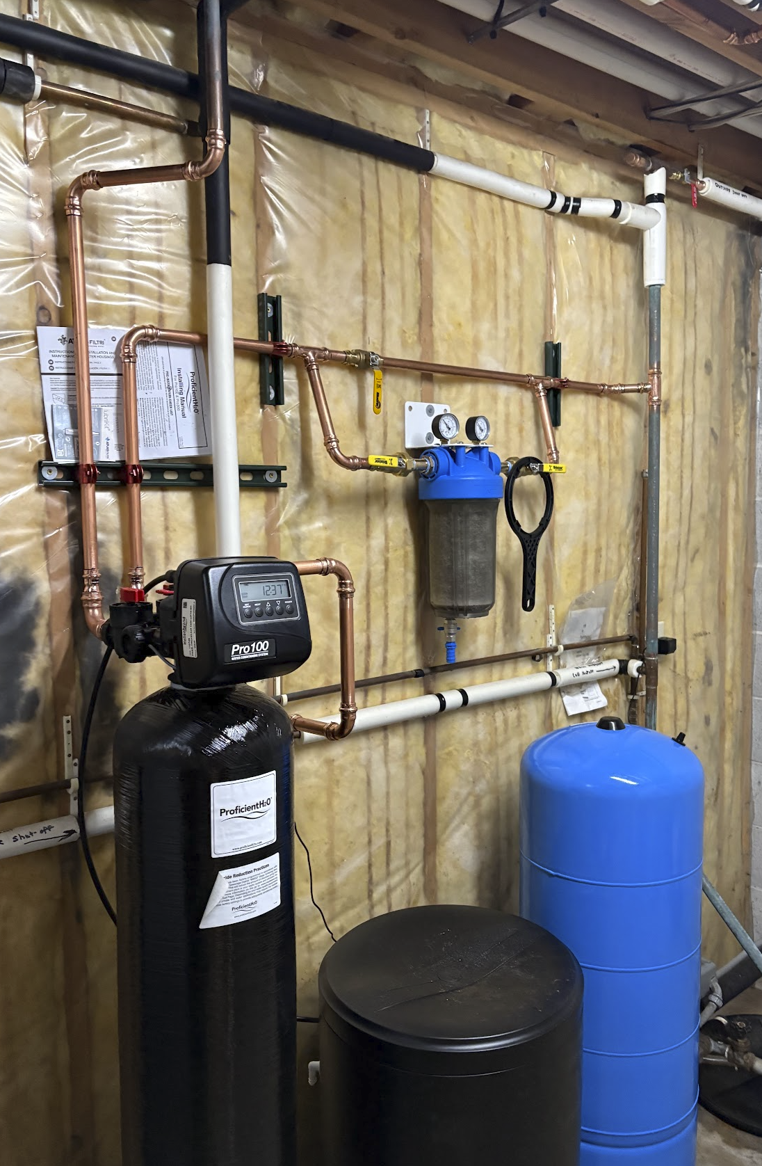Plumbing system with copper and PVC pipes, a water softener, and filtration units installed on a wooden wall in a utility room.