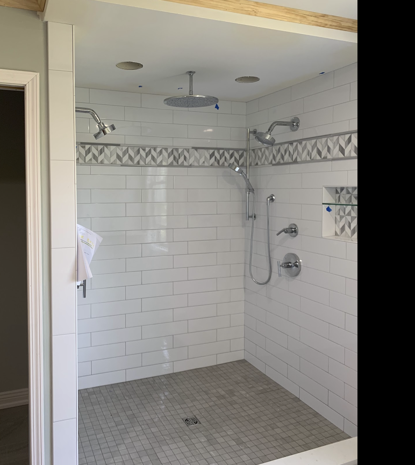 Newly installed walk-in shower with white subway tiles, decorative gray patterned border, and grab bars. Shower fixtures include a large rainfall showerhead, a handheld shower, and two adjuster control knobs.