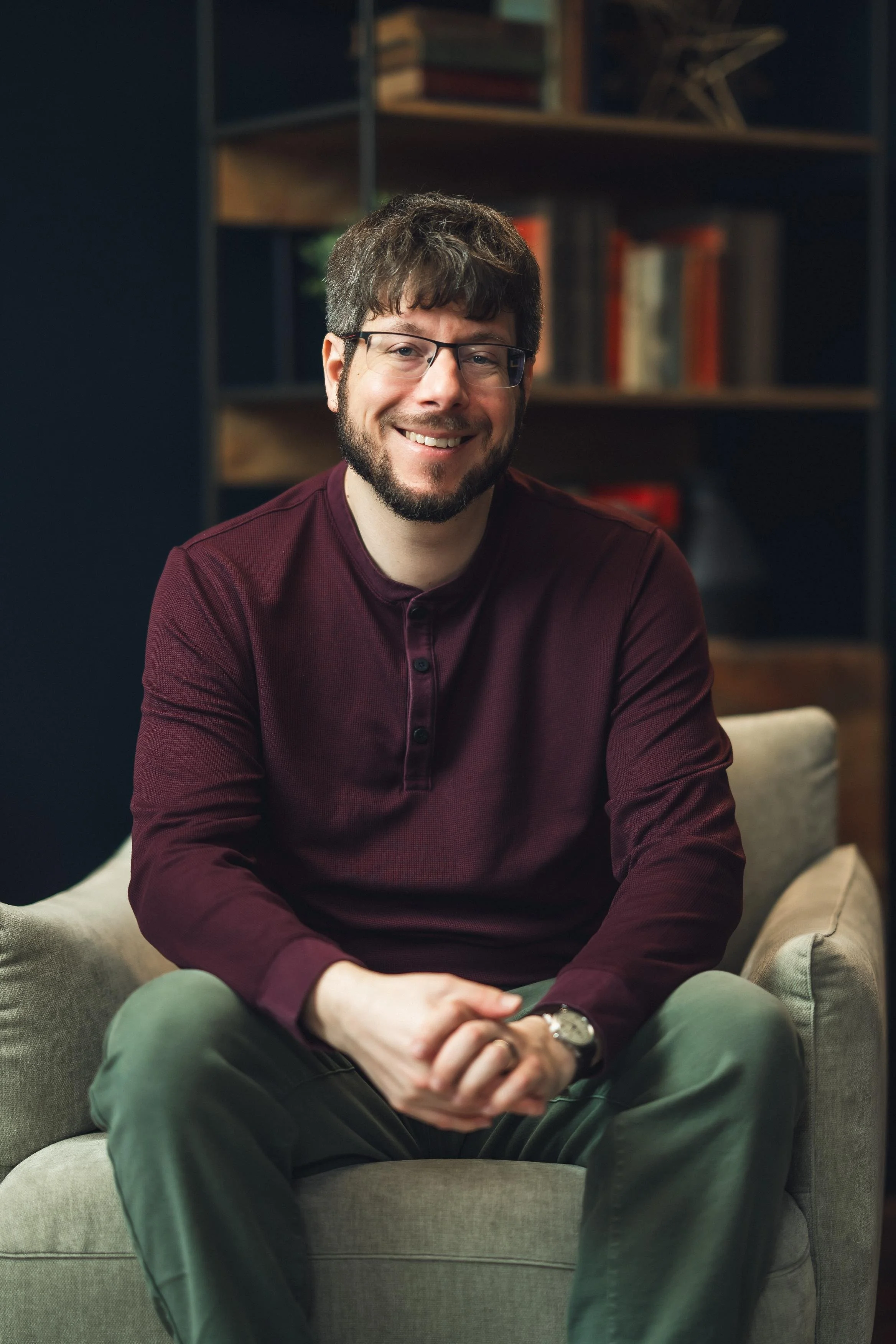 A smiling man with glasses and a beard wearing a burgundy shirt, sitting on a gray armchair in a cozy room with a bookshelf filled with books in the background.