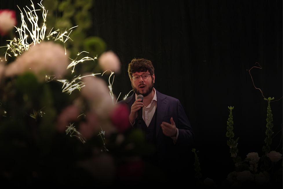 A man in a suit, wearing glasses, holding a microphone and speaking at an event with fireworks and floral decorations in the background.