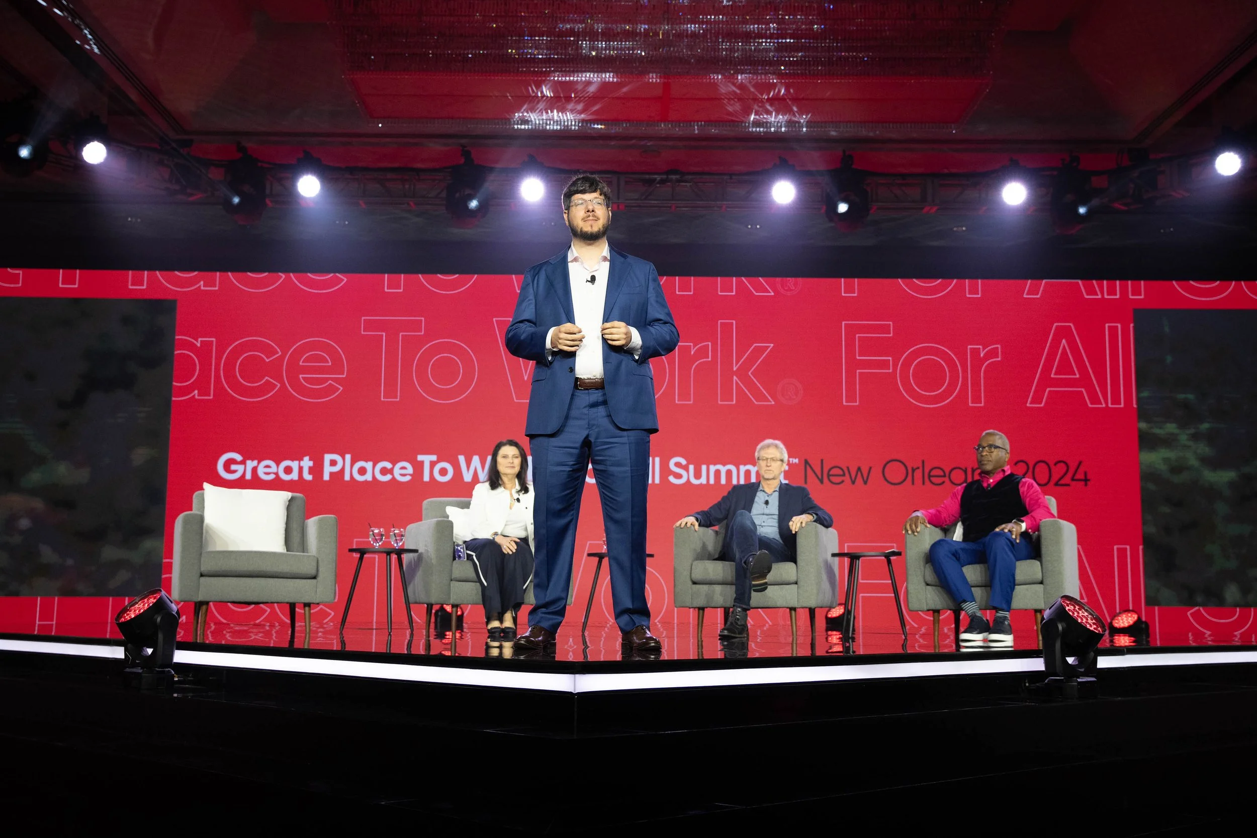 A man in a blue suit standing on a stage speaking at a conference with a panel seated behind him. The backdrop has large red text that says "Great Place to Work" and "All Summit". There are four panelists seated in chairs with small tables in front of them on the stage.