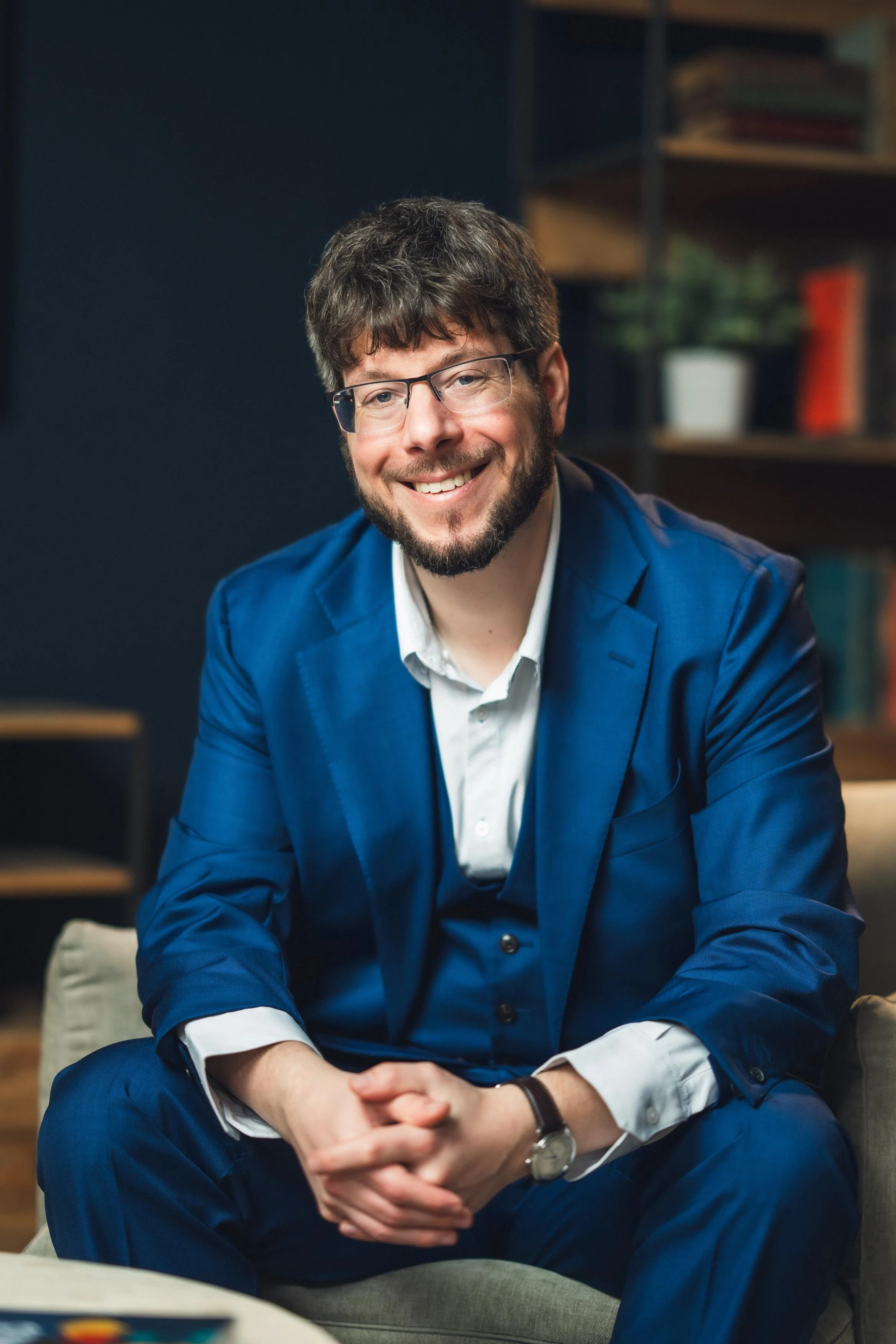 A man with glasses and a beard, dressed in a blue suit, smiling while sitting on a beige chair in an indoor setting with bookshelves in the background.