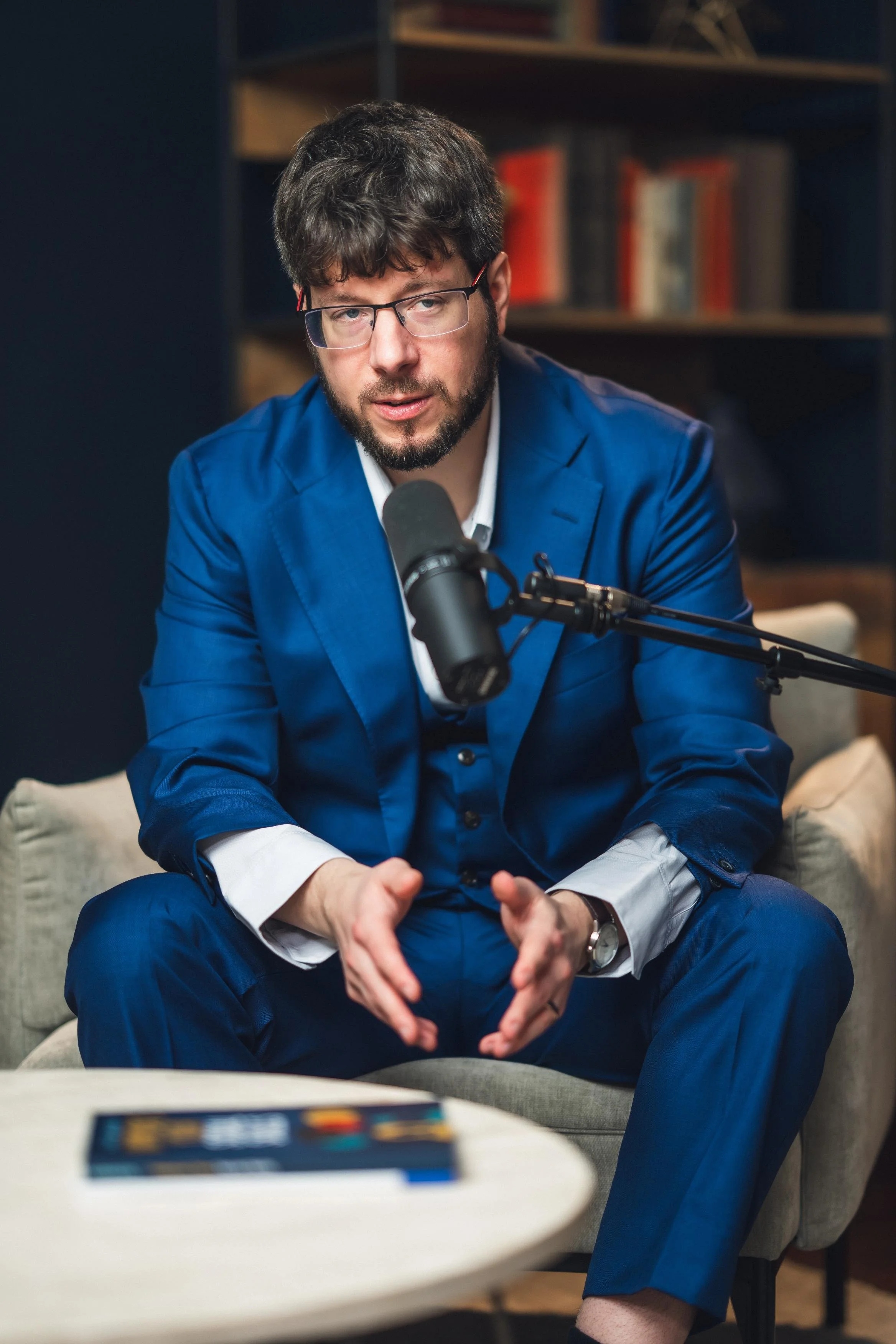 A man with dark hair, glasses, and a beard wearing a blue suit and white shirt, sitting on a gray couch in front of a microphone, engaging in a conversation or interview in a cozy room with bookshelves in the background.