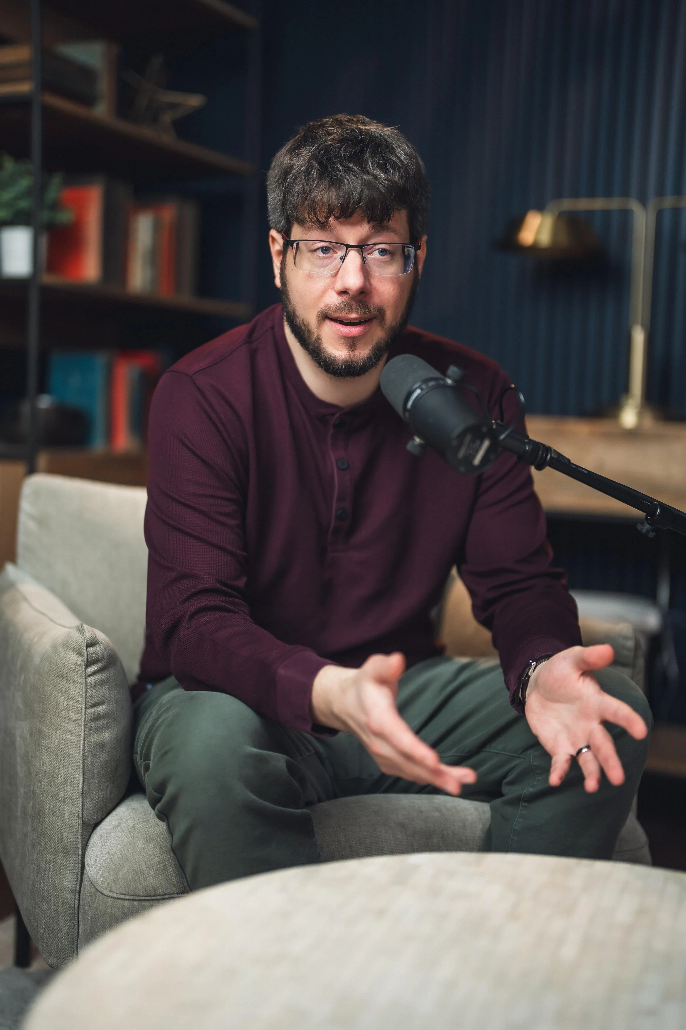 A man with glasses and a beard, wearing a maroon shirt, appears to be speaking into a microphone during an interview or podcast session. He is sitting on a light-colored couch in a room with dark blue walls, a bookshelf with books, and a gold desk lamp.